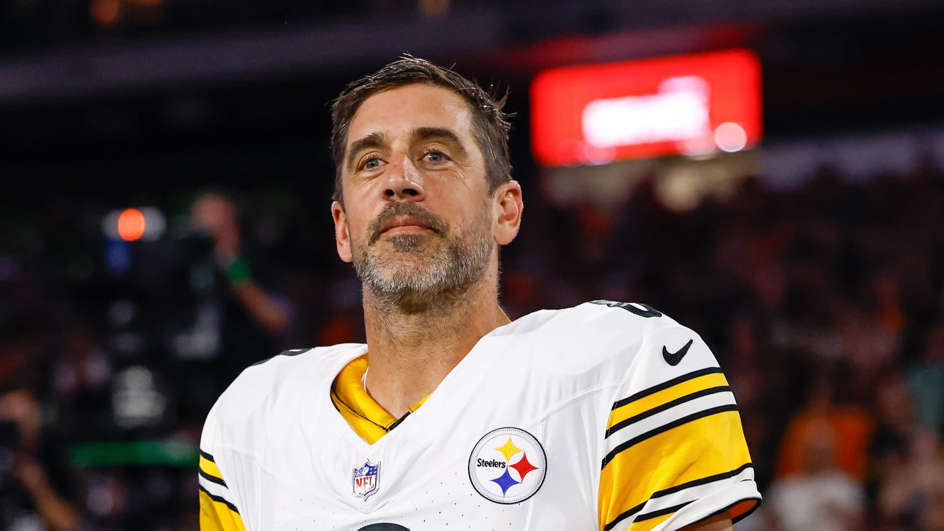 Aaron Rodgers a Pittsburgh Steelers jersey stands on a football field, looking upward with a focused expression.