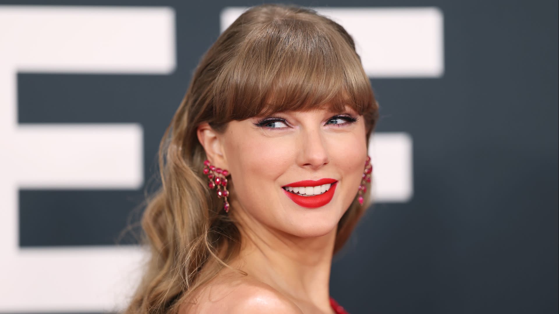 Taylor Swift smiling, wearing red lipstick and earrings, with wavy hair and bangs, at a formal event.
