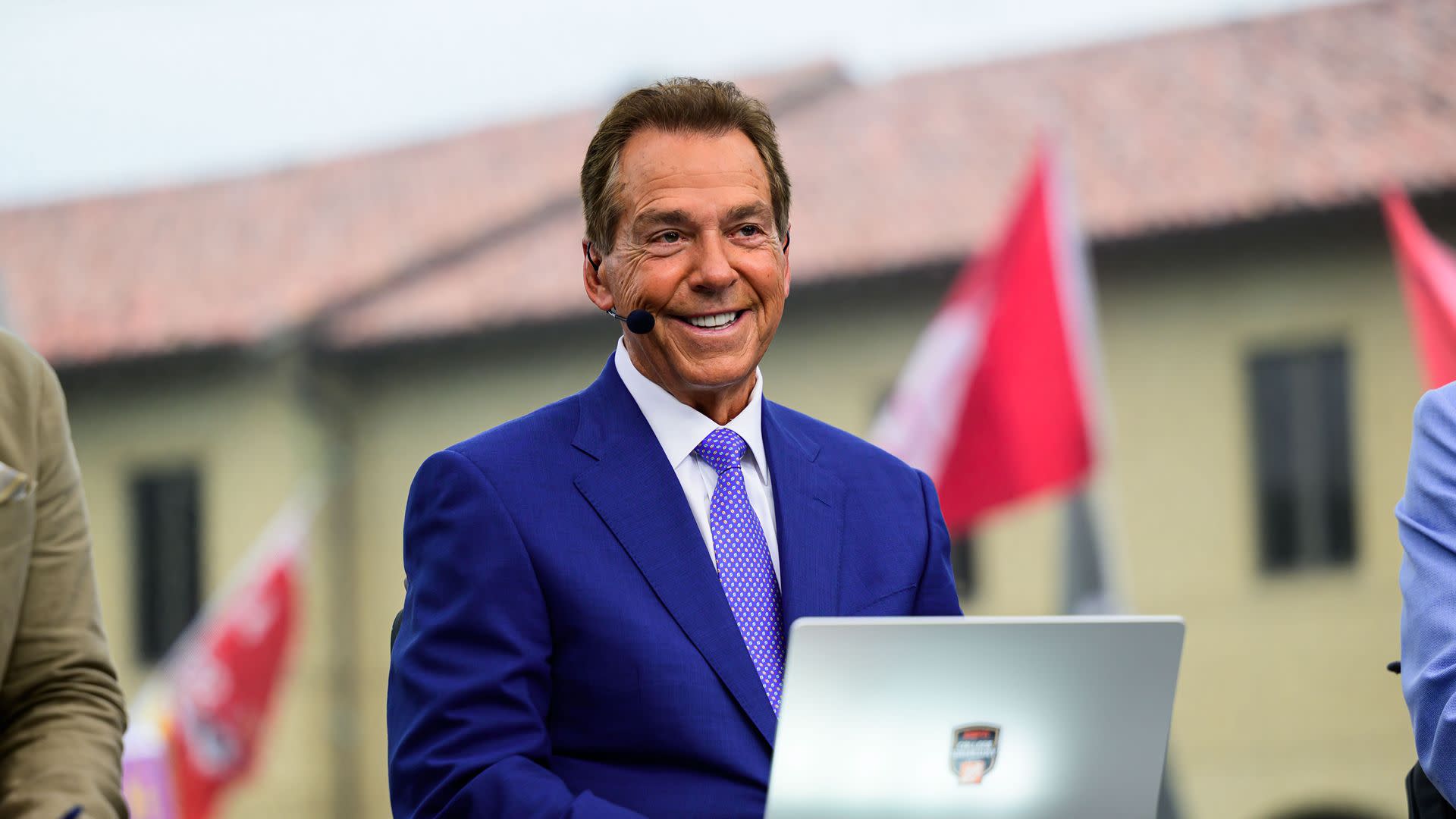 A man in a blue suit with a microphone headset, smiling, seated outdoors with a laptop. Flags and a building are in the background.