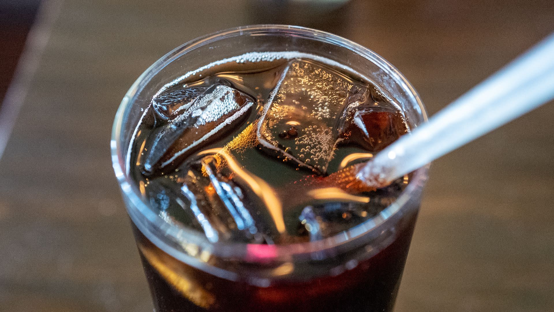 A glass of soda with ice cubes and a straw on a wooden table.