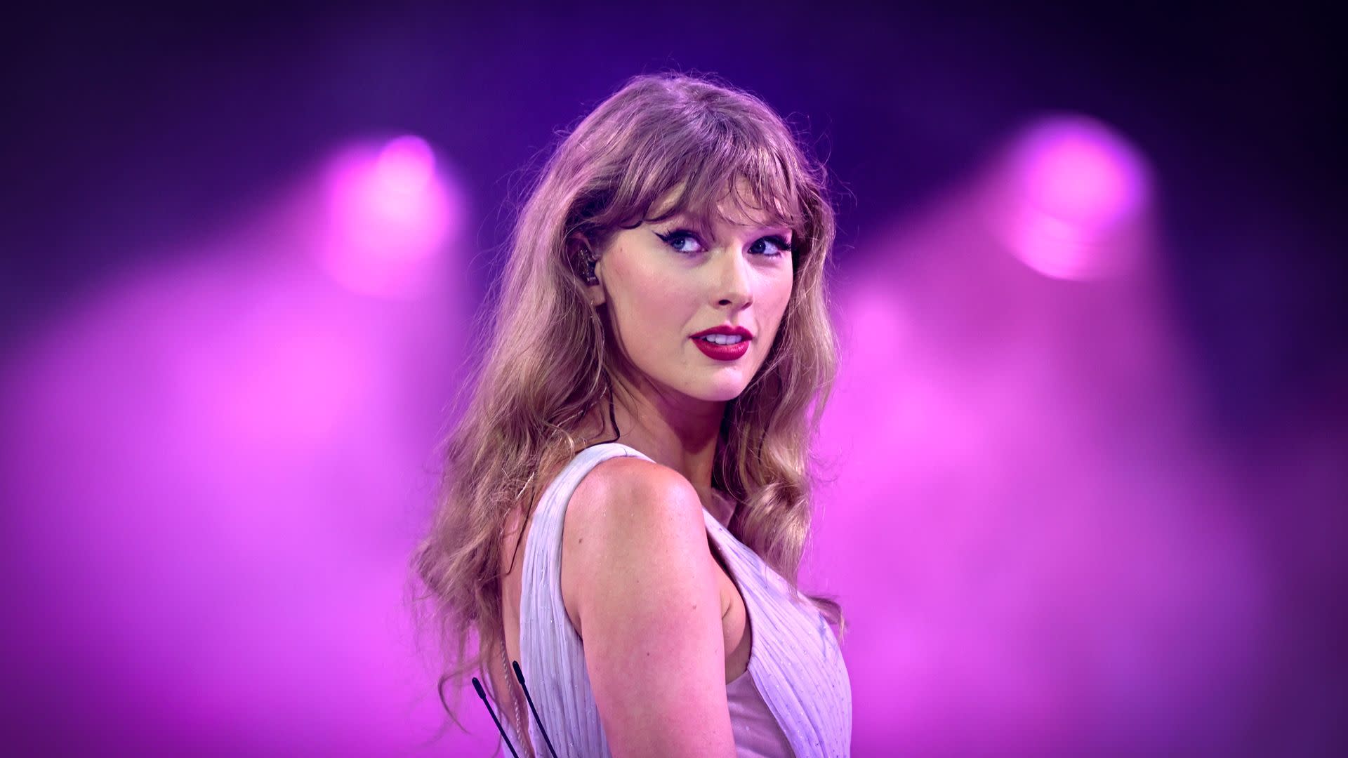 Taylor Swift performing on stage with a purple backdrop, wearing a light-colored dress and looking to the side.