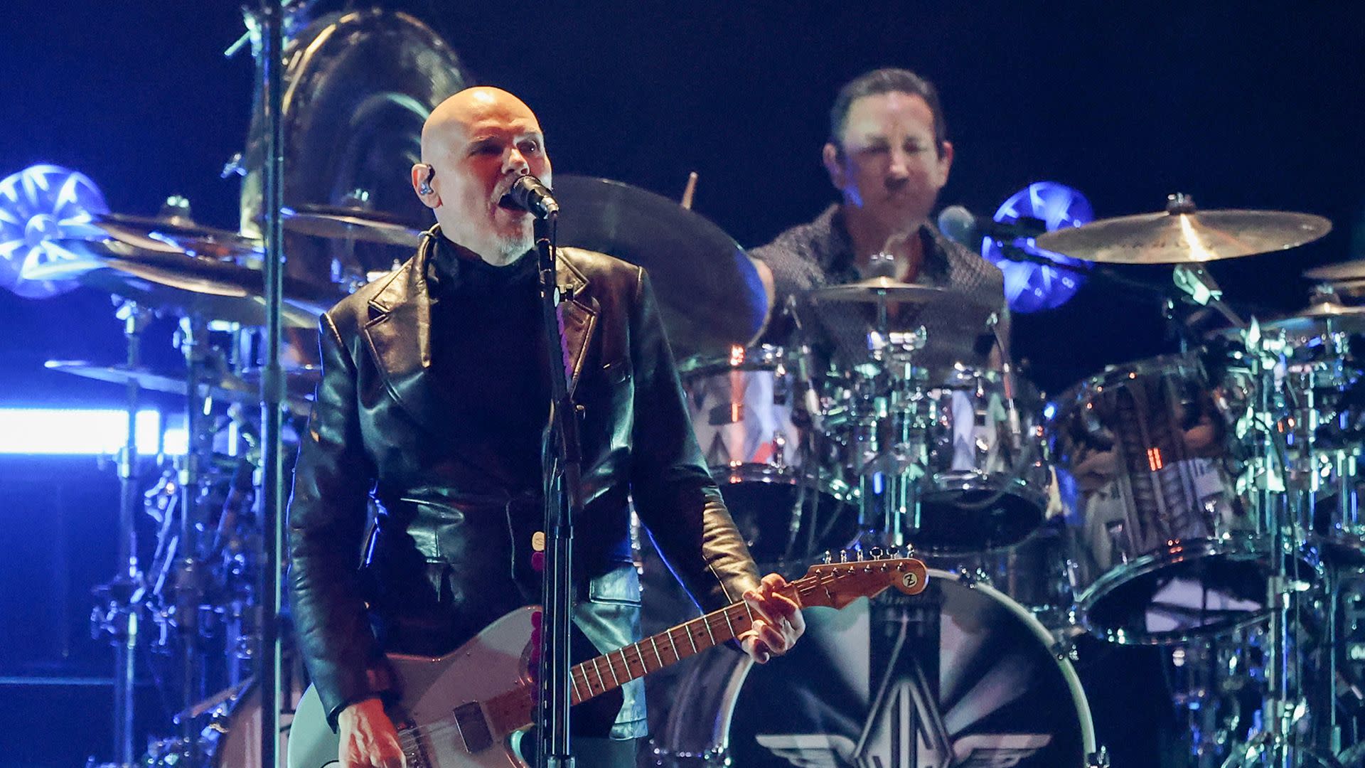Billy Corgan performing on stage with a guitarist and a drummer in the background.