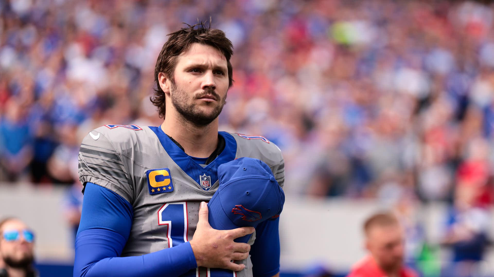 A football player in a gray uniform holds a cap to his chest, standing on a field with a blurred crowd in the background.