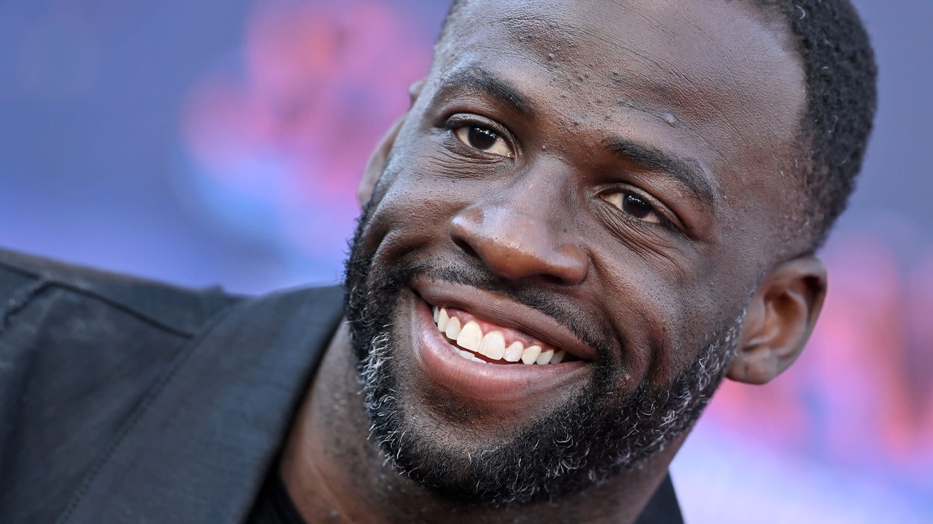 A smiling man with a beard, wearing a black jacket, against a blurred colorful background.