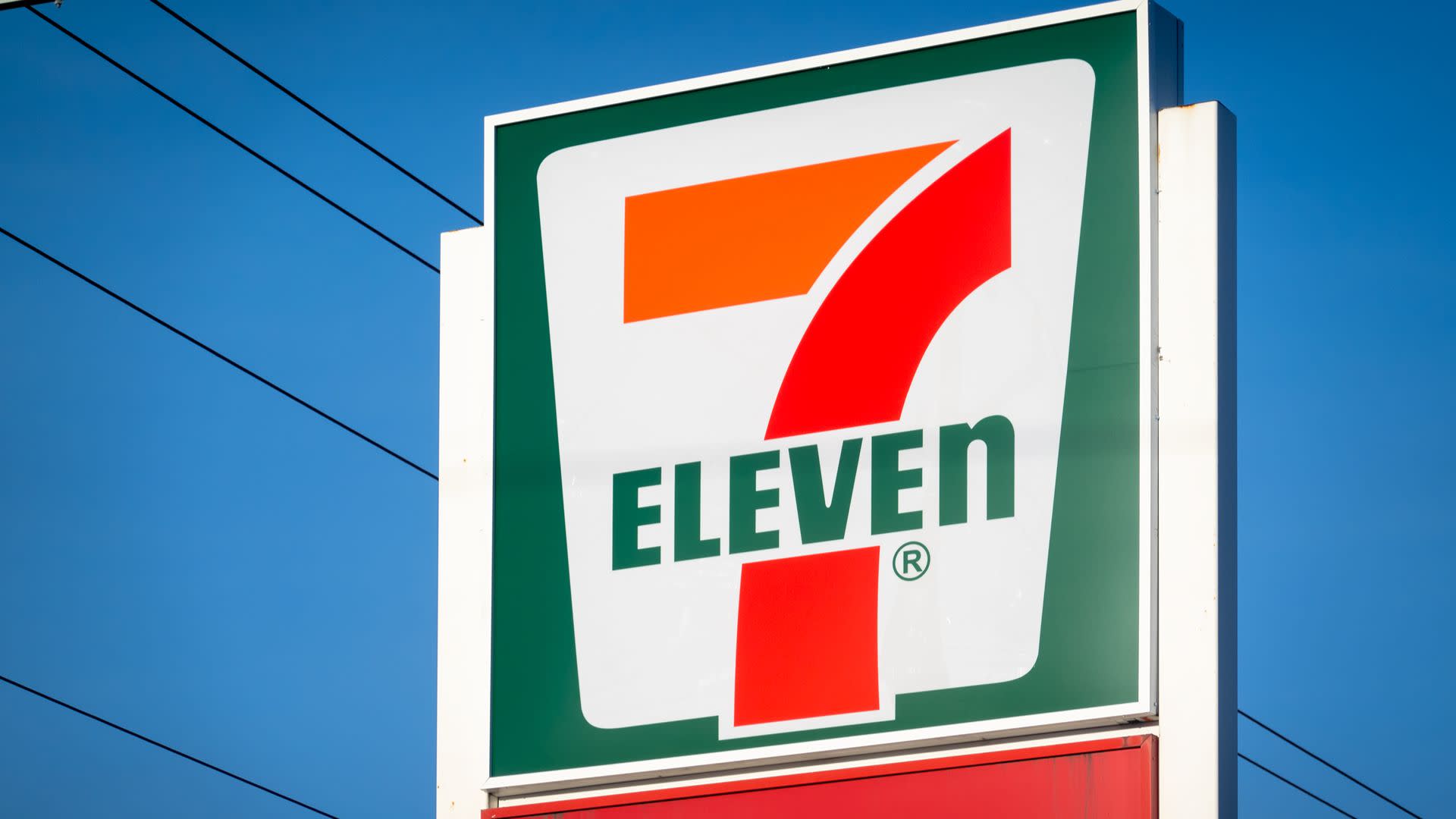 A 7-Eleven store sign against a clear blue sky, featuring the iconic red and green logo with the number 7 prominently displayed.