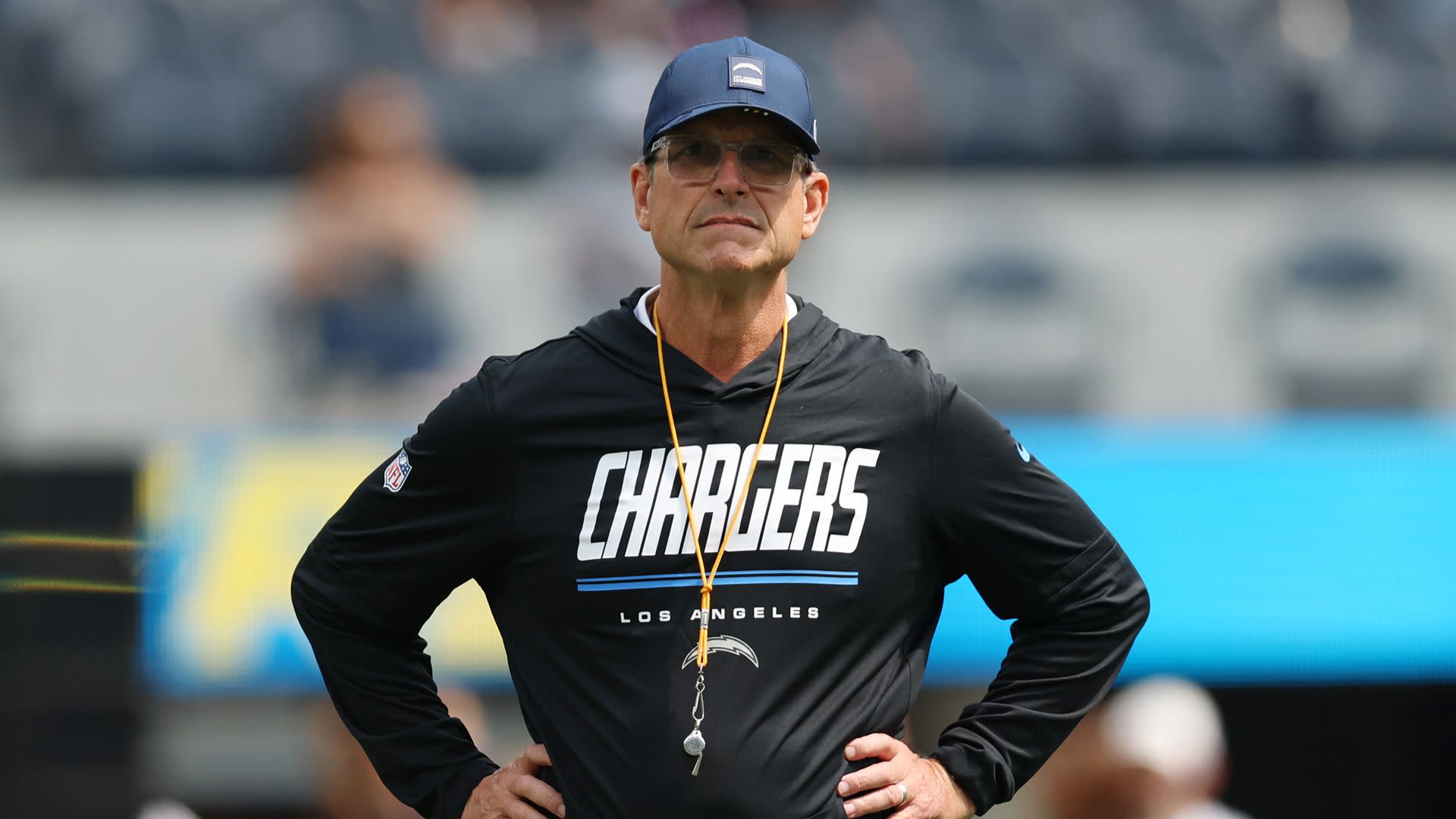 A man wearing a Los Angeles Chargers sweatshirt and cap, standing with hands on hips in a stadium.