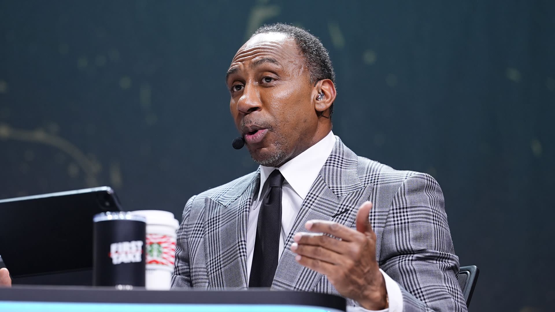 Stephen A. Smith speaking passionately on a set, wearing a plaid suit, with a headset microphone.