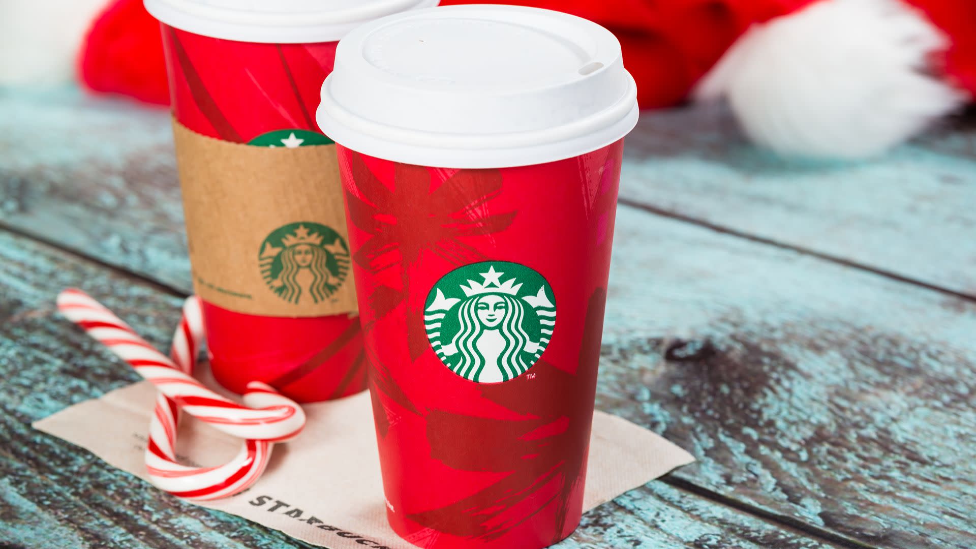 Two red Starbucks holiday cups with white lids on a wooden surface, next to candy canes and a Santa hat.