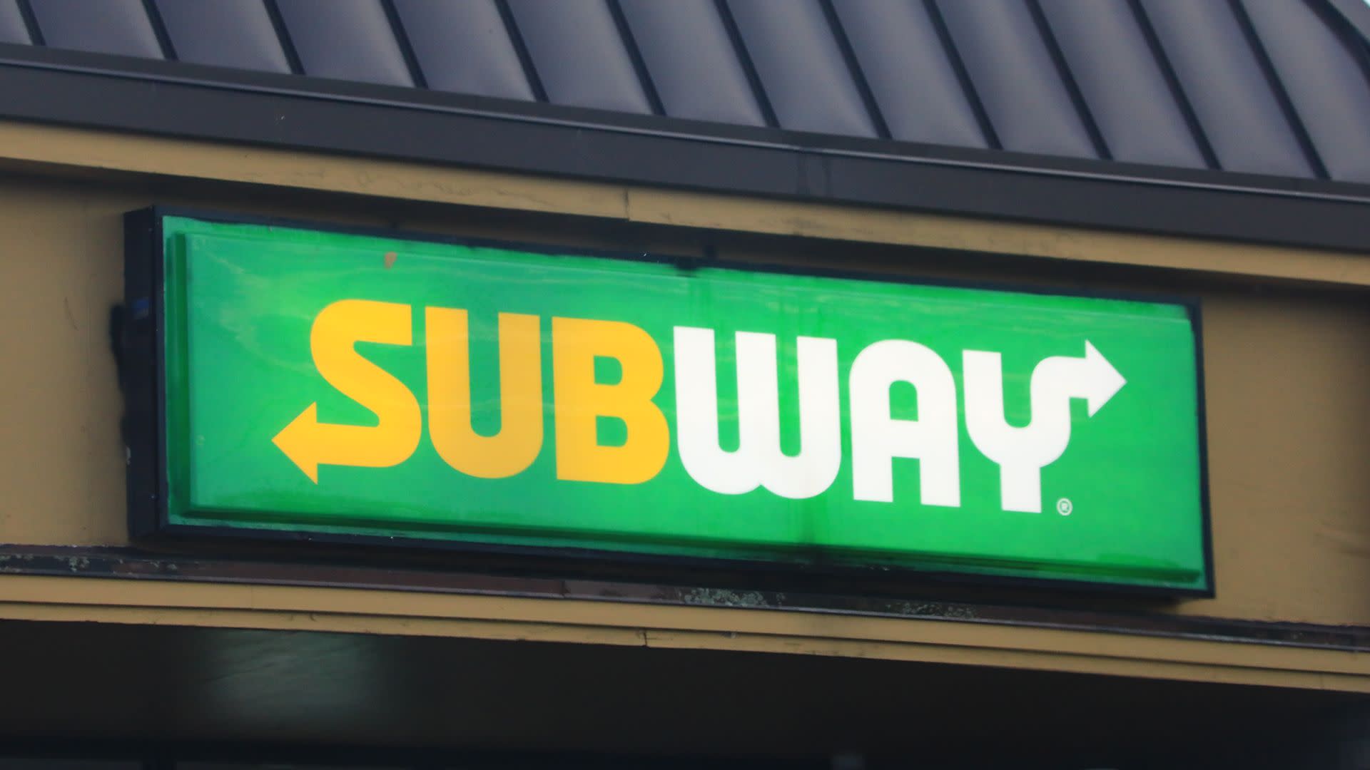 A Subway restaurant sign with a green background and yellow and white text, featuring arrows on both ends.