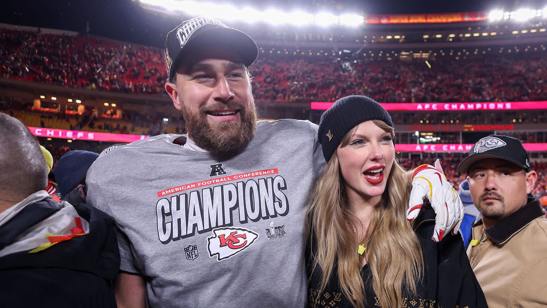 Travis Kelce in a "Champions" shirt and Taylor Swift in a black beanie celebrate on a football field.