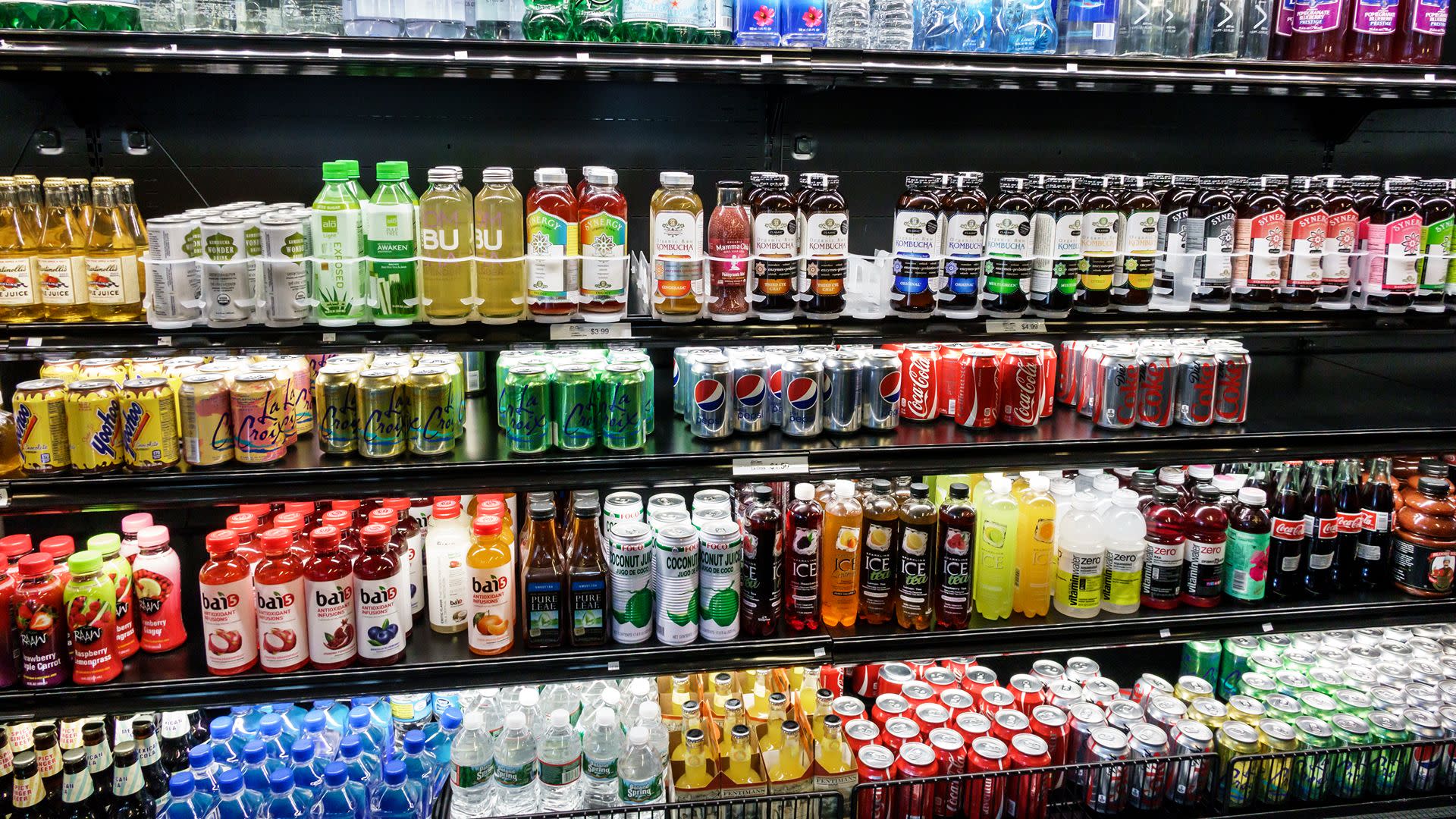 A refrigerated display filled with various beverages, including soda cans, bottled juices, and sparkling water, in a store setting.