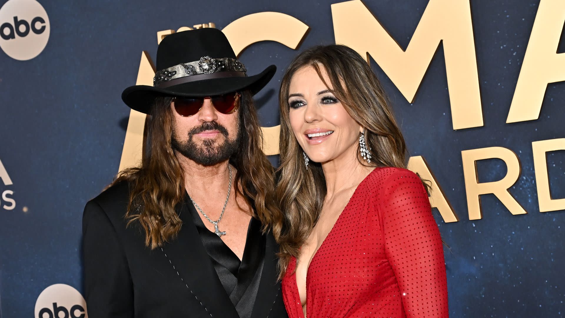 Billy Ray Cyrus in a black hat and sunglasses, with Elizabeth Hurley in a red dress, smiling at an awards event.