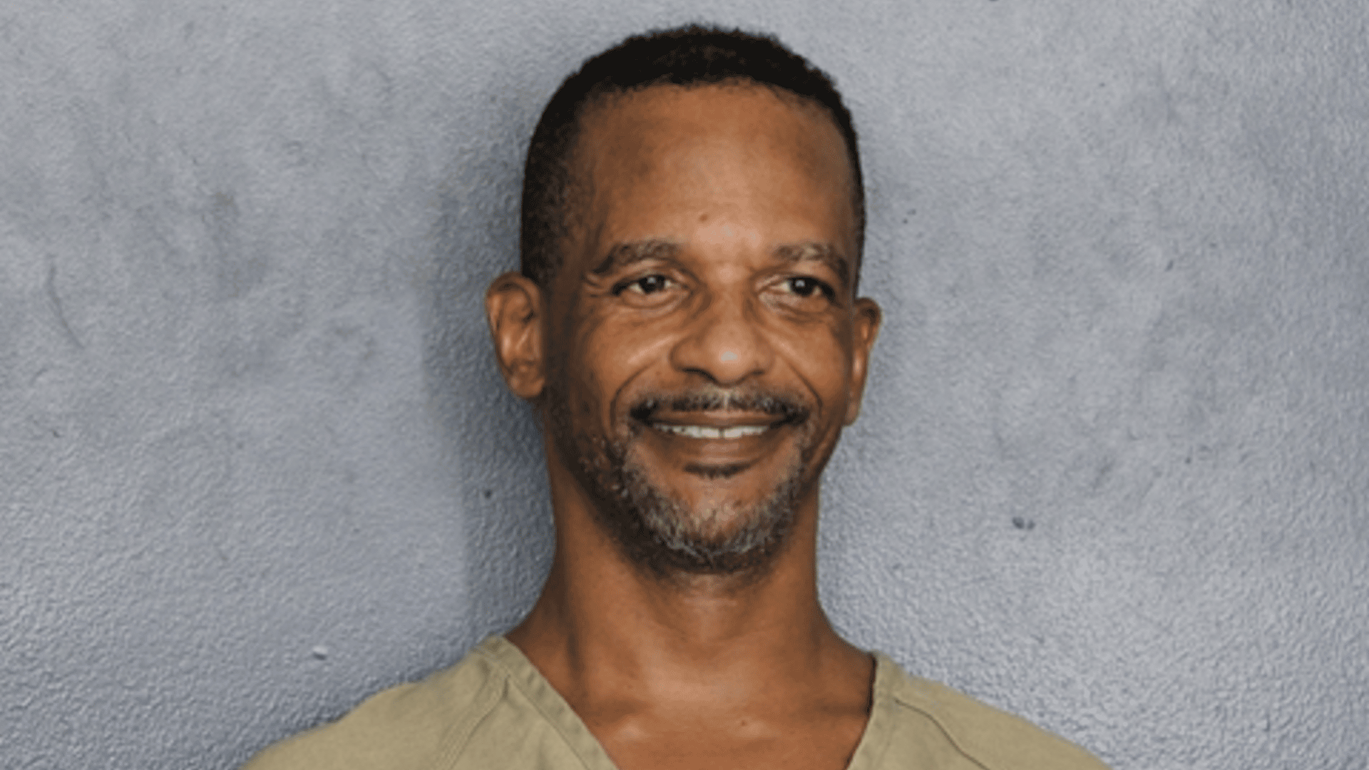 Charleston White with short hair and a beard, wearing a beige shirt, smiling against a gray background.