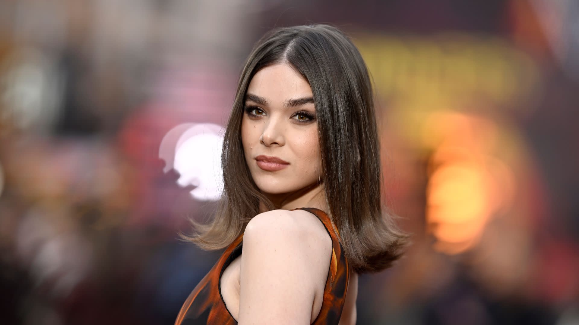 Hailee Steinfeld in a patterned dress poses confidently, with a blurred background of lights.