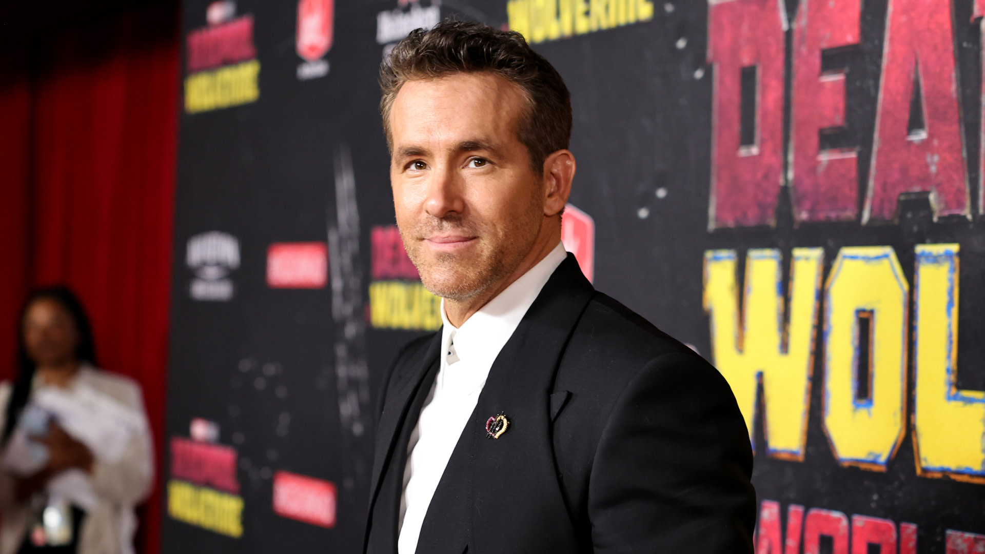 Ryan Reynolds at a red carpet event, wearing a black suit and white shirt, with "Deadpool" and "Wolverine" signage in the background.