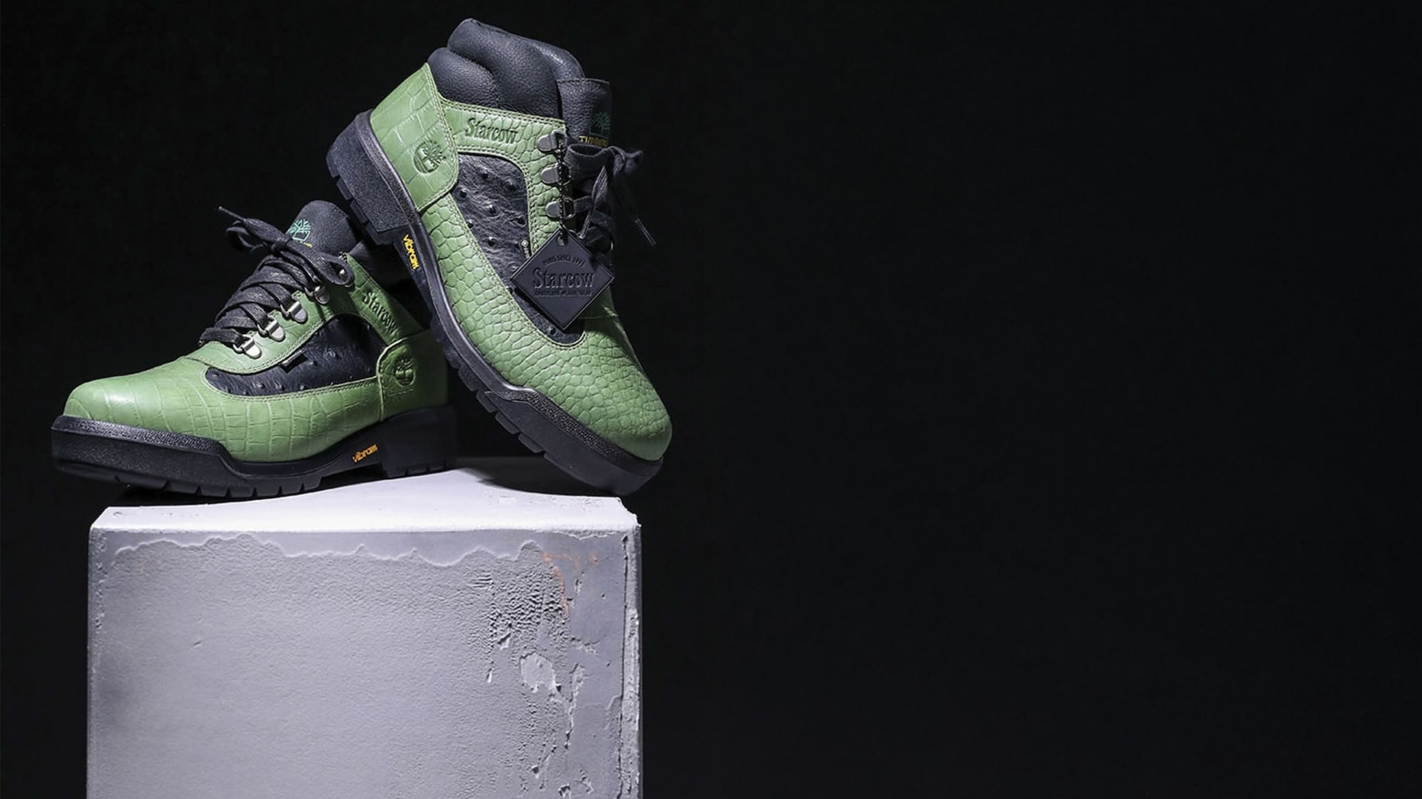 A pair of green and black sneakers with a textured pattern, displayed on a concrete pedestal against a dark background.