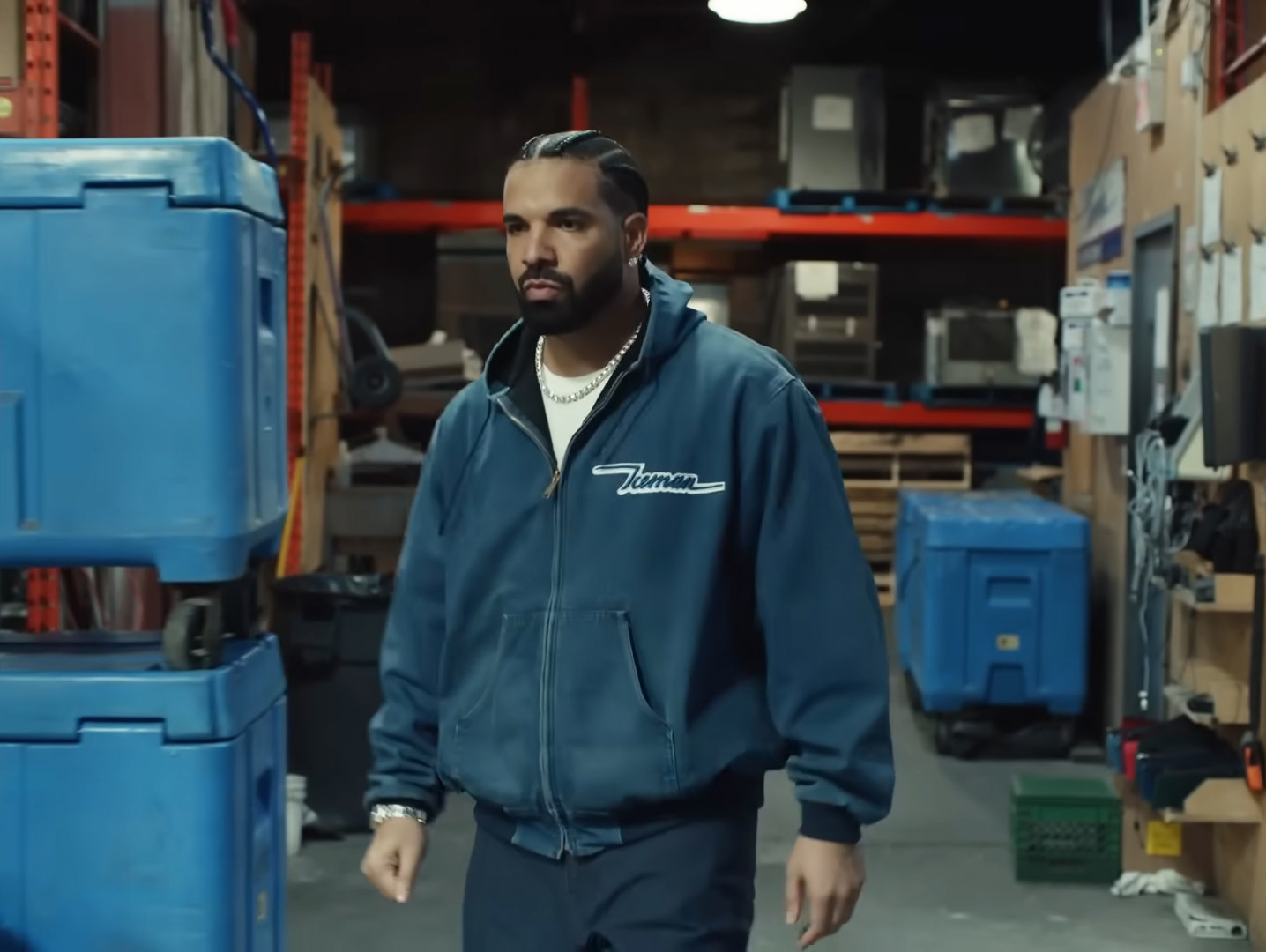 Drake in a warehouse, wearing a blue jacket with "Truman" on it, surrounded by blue bins and shelves.