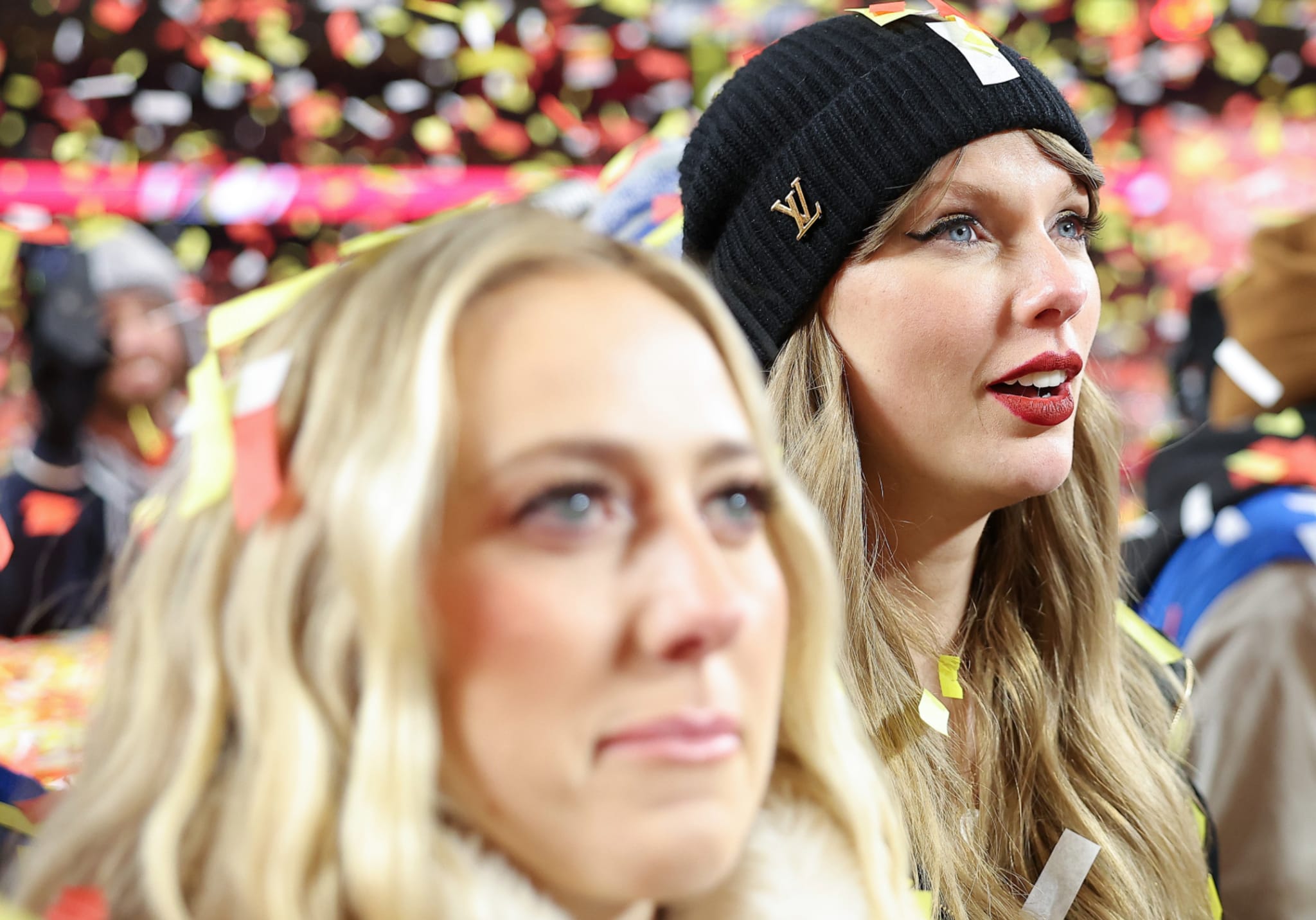Two women in a crowd with confetti falling. One wears a black beanie and red lipstick. The other has blonde hair.