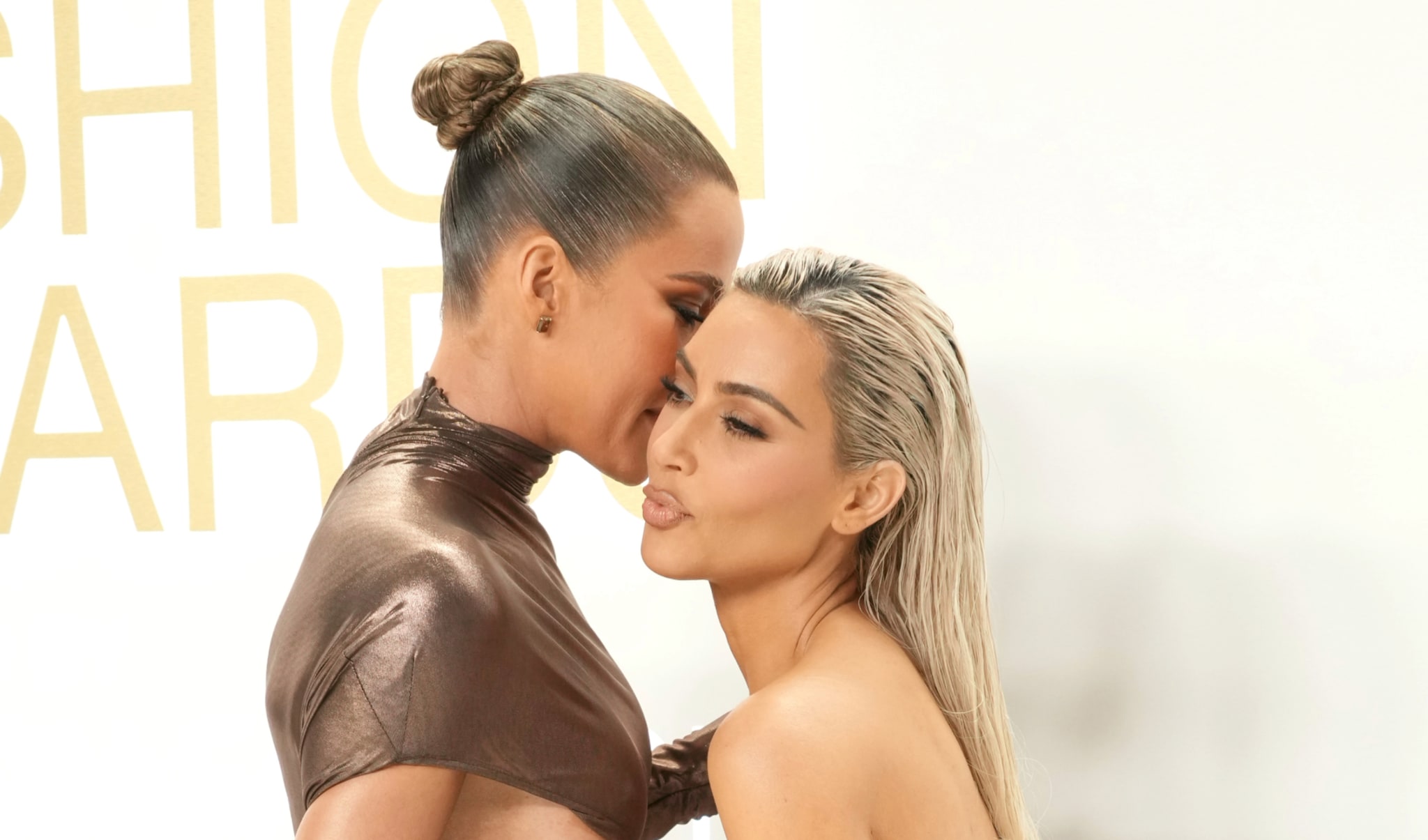 Two women embracing at an event, one in a metallic dress with a bun, the other with long, blonde hair.