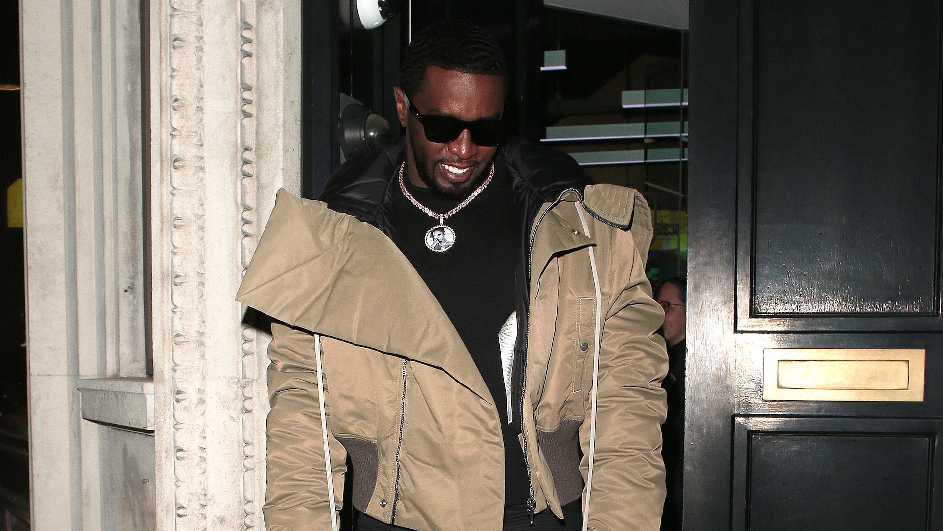 Diddy wearing sunglasses and a tan jacket with a black shirt underneath, smiling as he exits a building.