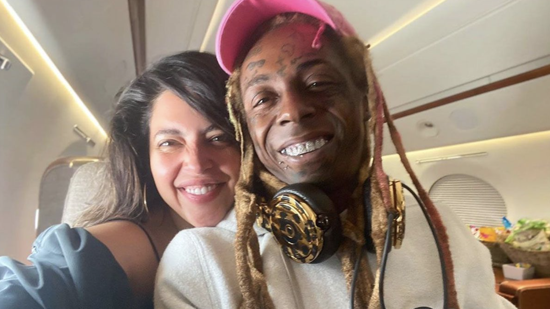 Lil Wayne, wearing a pink cap and headphones, smiles alongside Denise Bidot on a private jet.