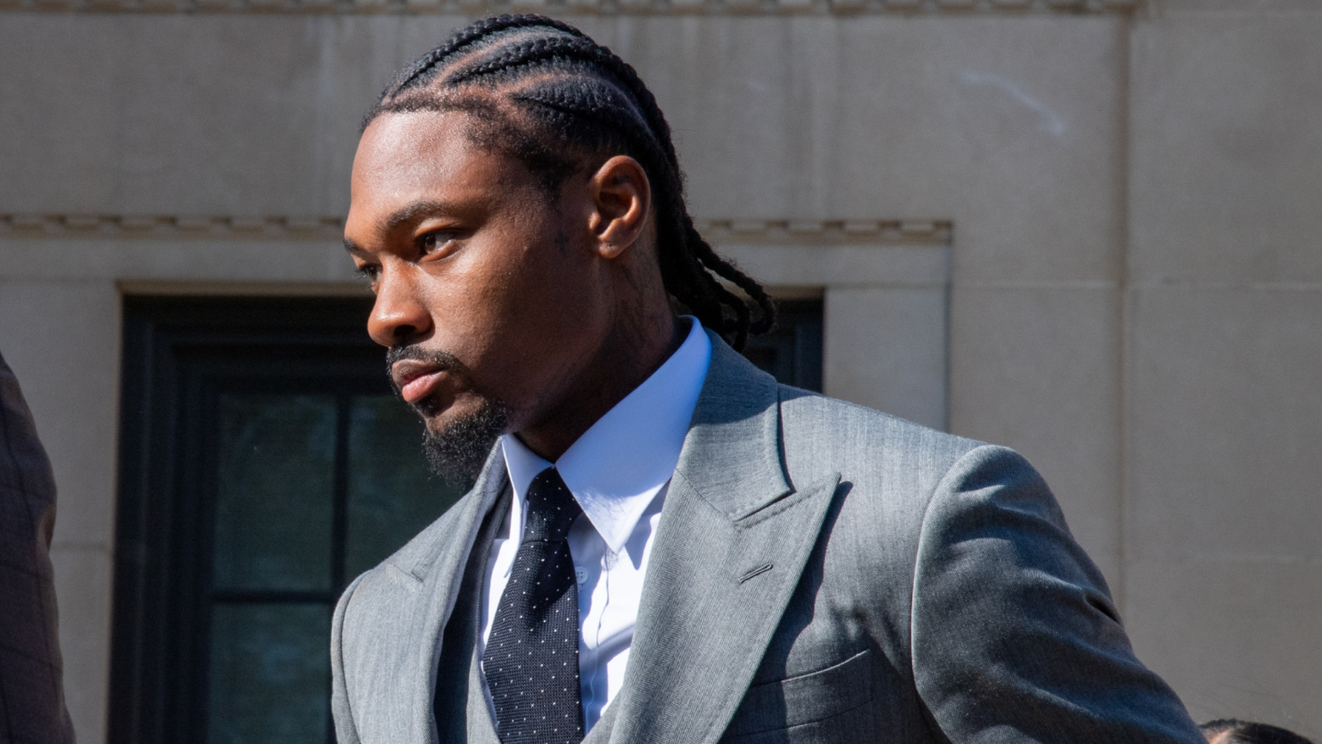 Stefon Diggs in a gray suit with cornrows, standing outside a building, looking serious.