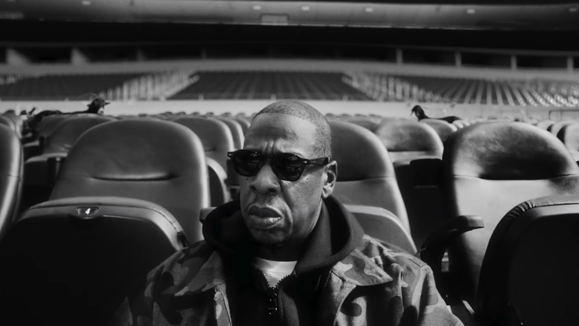Jay-Z wearing sunglasses and a jacket sits alone in a stadium with empty seats around him, in a black and white image.