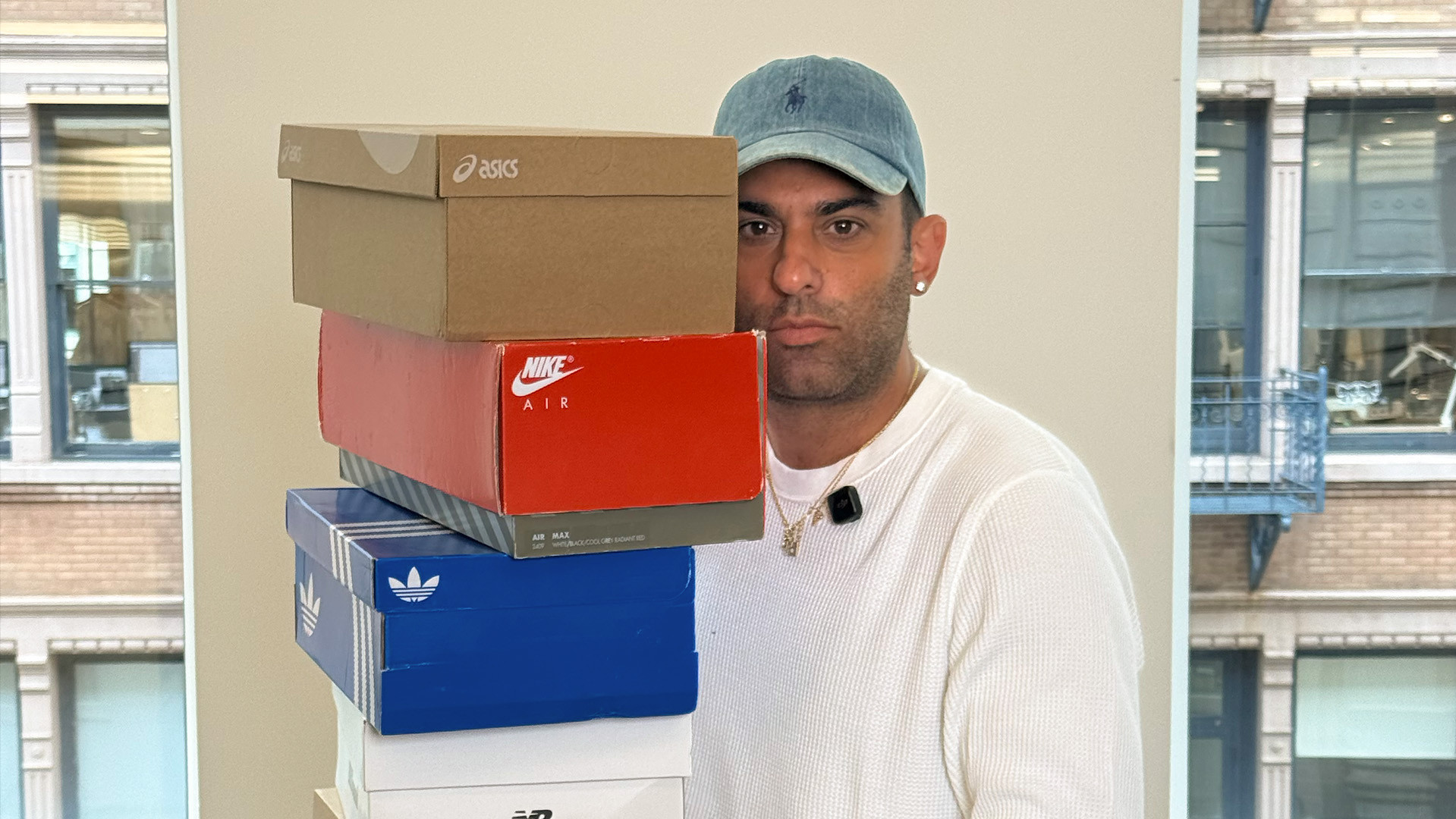 A person in a white shirt and blue cap holds a stack of shoeboxes from brands like Nike, Adidas, and Asics, against an urban window backdrop.