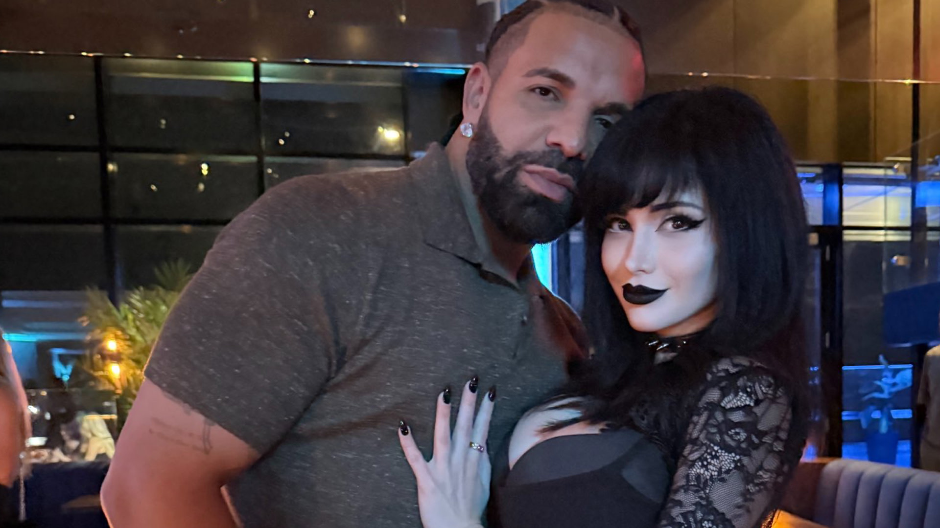 Drake in a grey shirt and a woman in black lace clothing with dark makeup pose together in a dimly lit room.