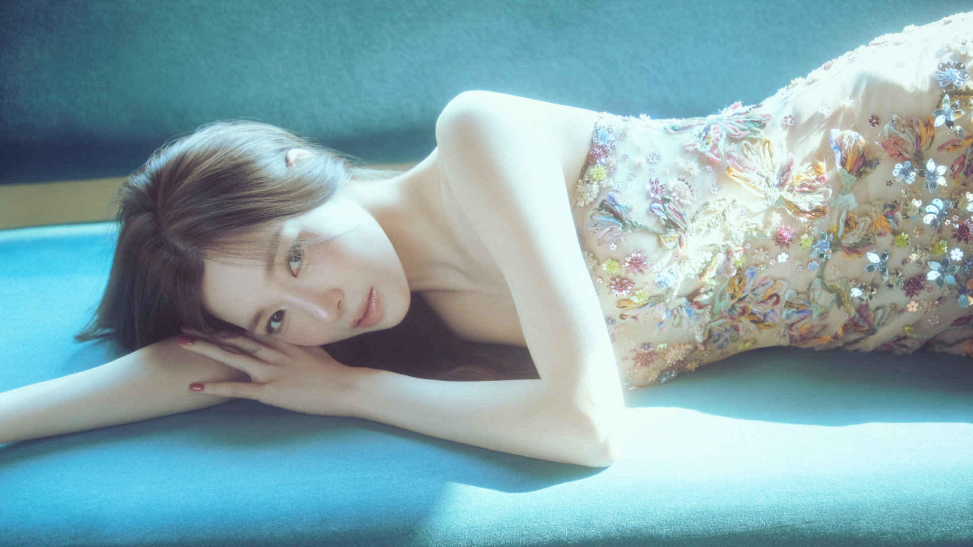 Tiffany Young in a floral embroidered dress lies on a teal surface, bathed in soft light.