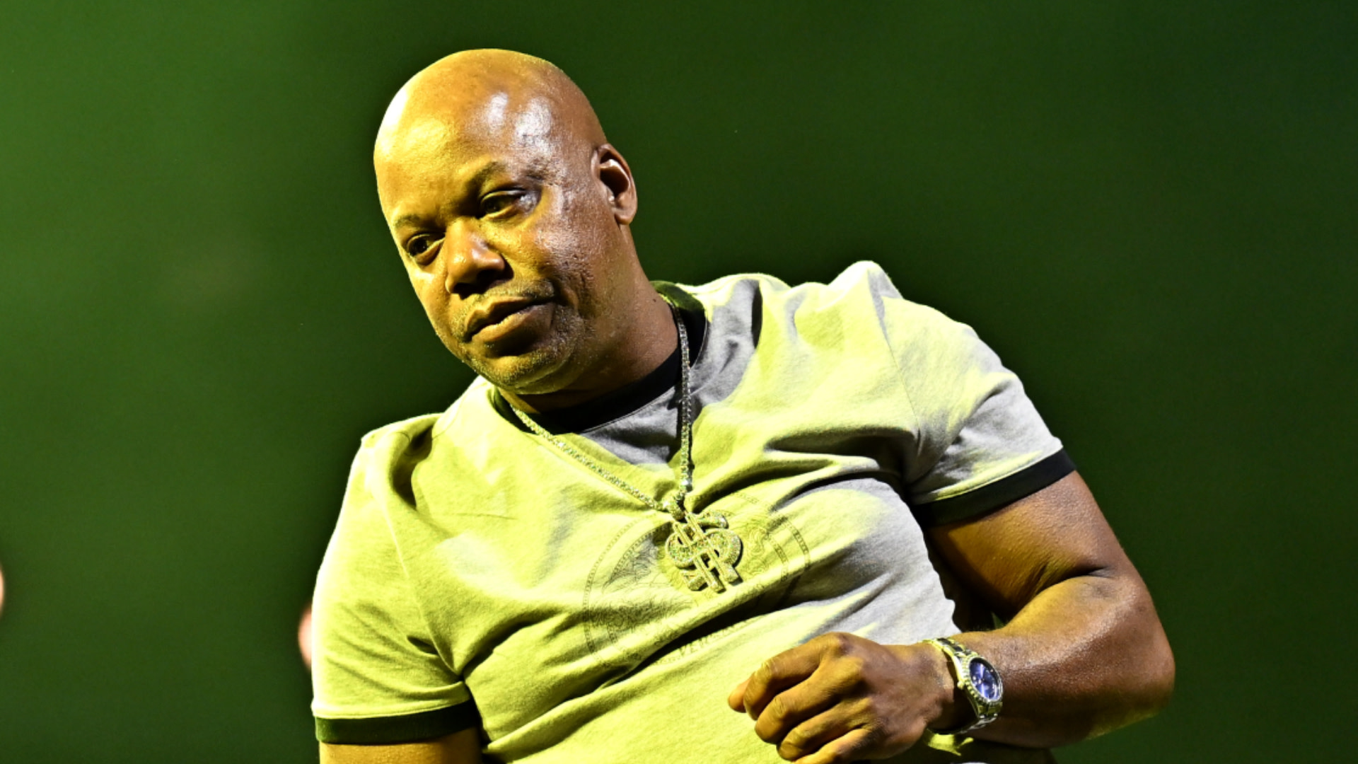Too $hort in a light-colored shirt and chain necklace stands against a green background.