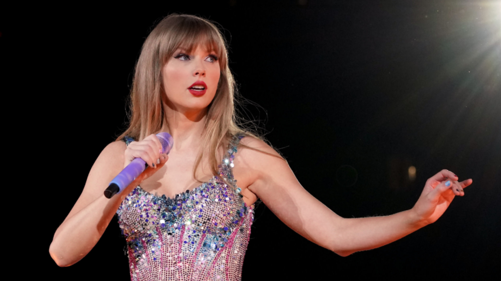 Taylor Swift performing on stage in a sparkling dress, holding a purple microphone, with a spotlight shining on her.