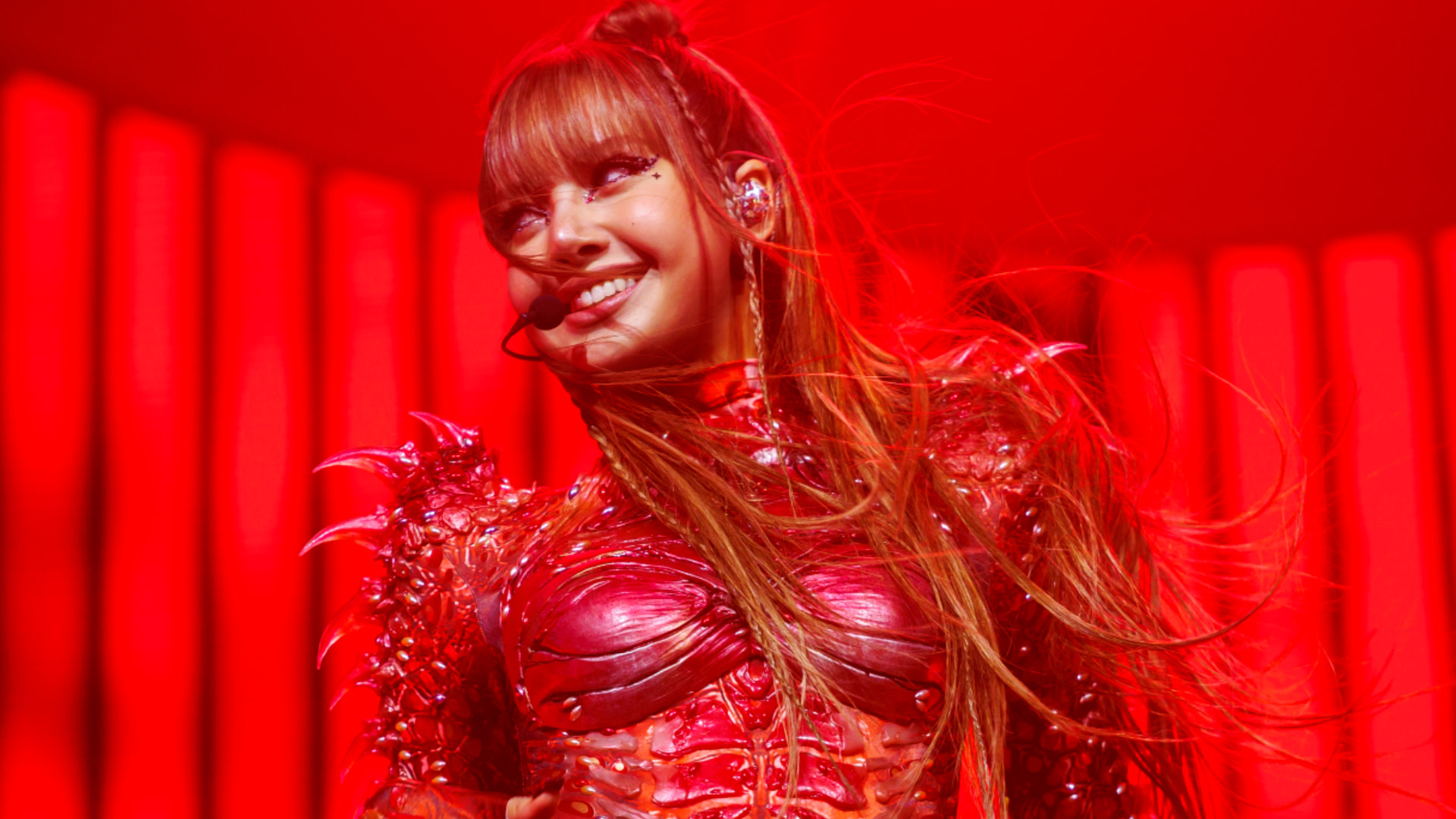 LISA in a red costume with long hair and a headset microphone, smiling on stage with a red background.
