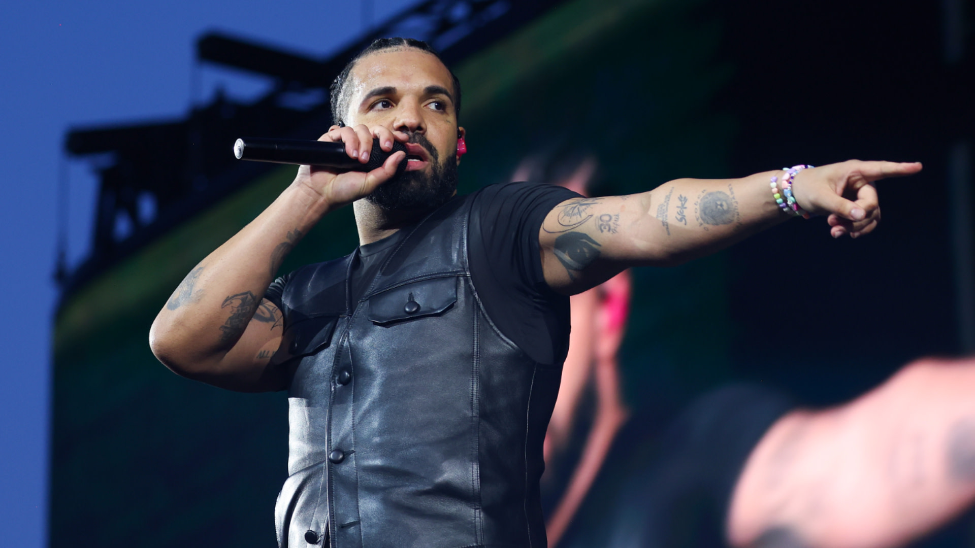 Drake performing on stage, wearing a black leather vest and holding a microphone, pointing outward with tattoos visible on his arms.