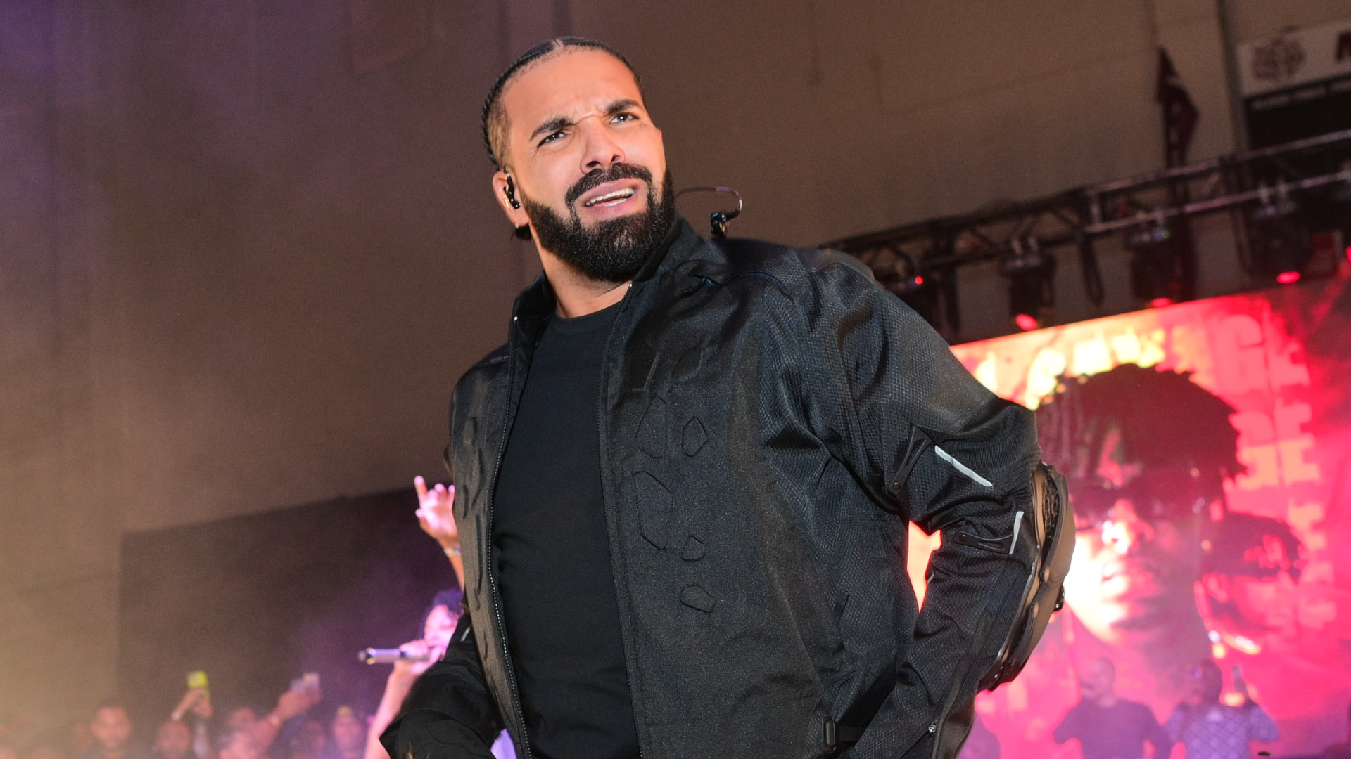 Drake performing on stage, wearing a black jacket, with vibrant lights and a crowd in the background.