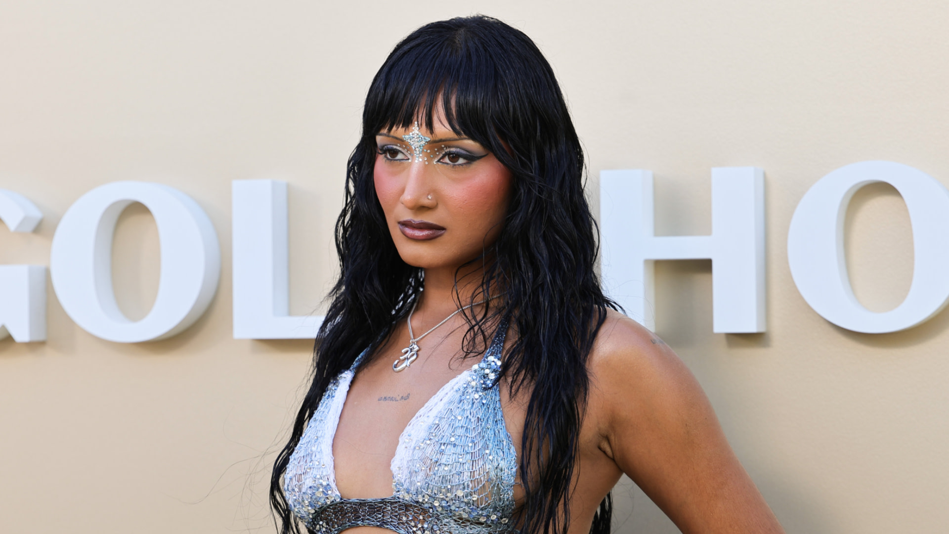 Rhea Raj with long dark hair and glittery makeup wearing a silver outfit, posing in front of a light-colored backdrop.