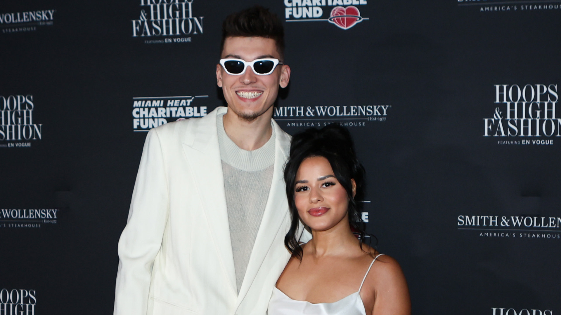 Tyler Herro in a white suit and sunglasses poses with Katya Elise Henry in a white dress at an event.