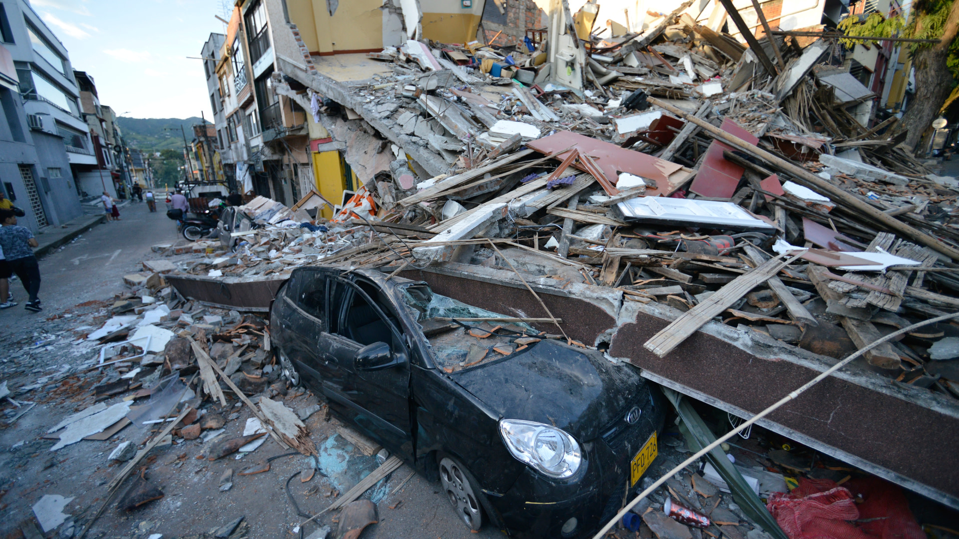 A black car crushed under debris from a collapsed building on a street, with surrounding rubble and damage visible. 
