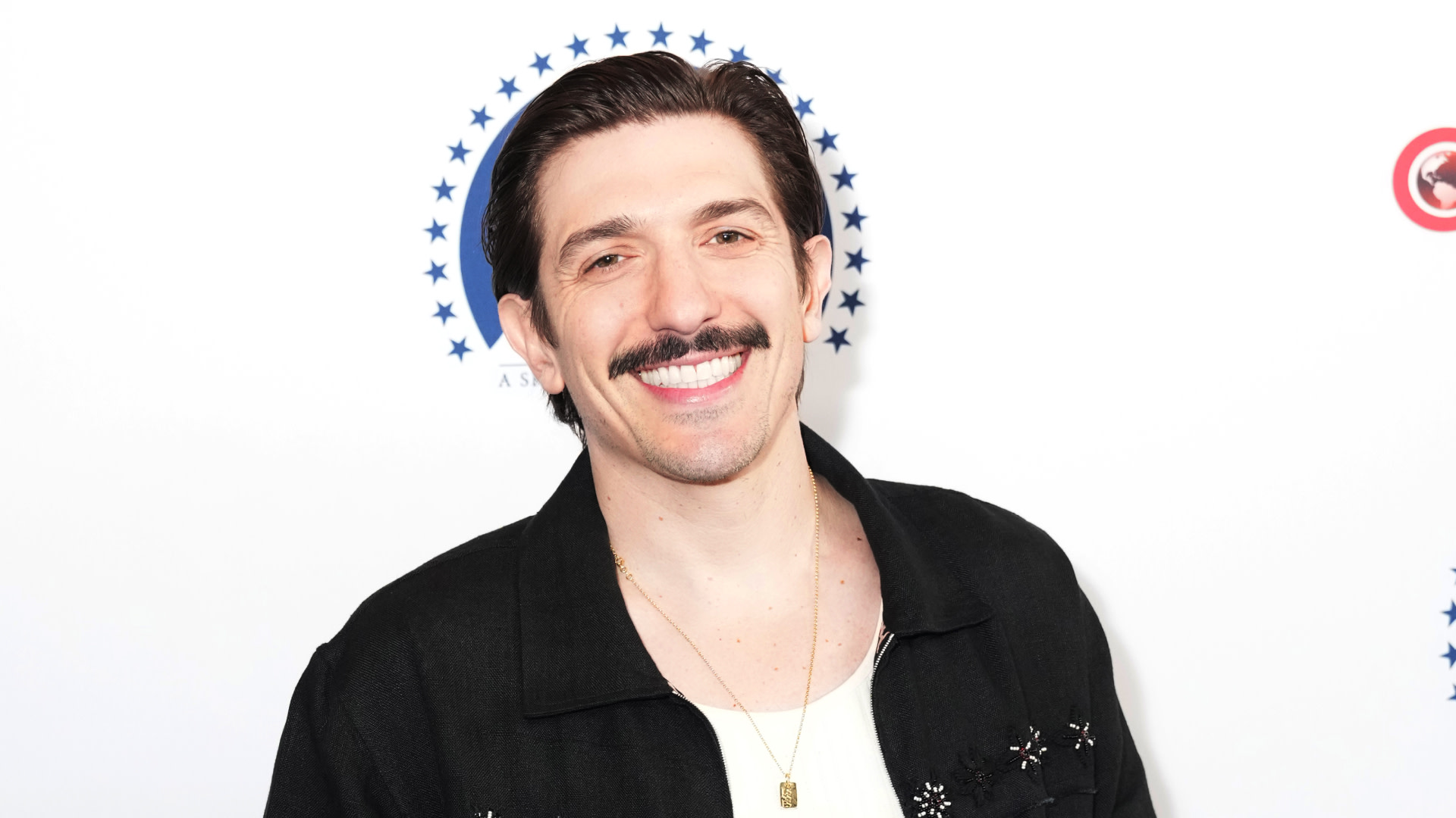 Andrew Schulz with a mustache smiling, wearing a black jacket and white shirt, standing against a white background with a circular star pattern.