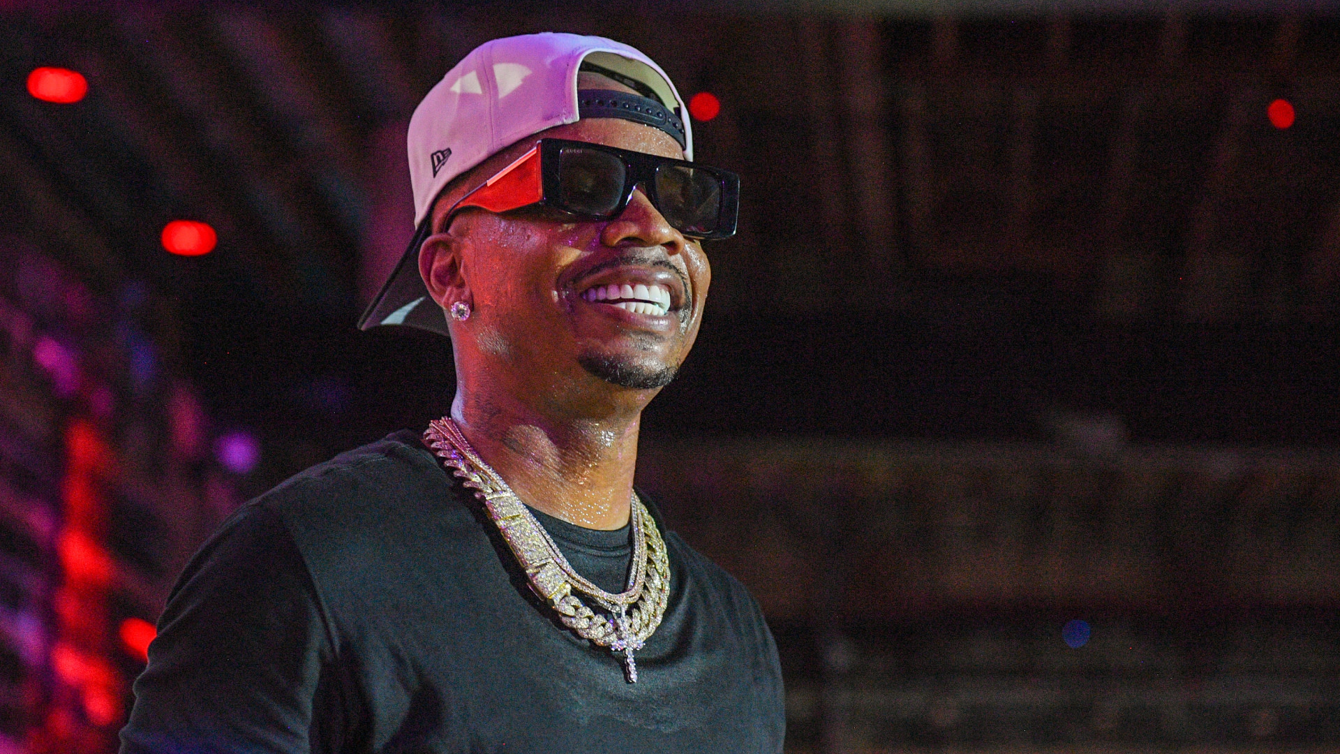 Plies wearing sunglasses, a backward cap, and a chain necklace smiles on stage under colorful lights.