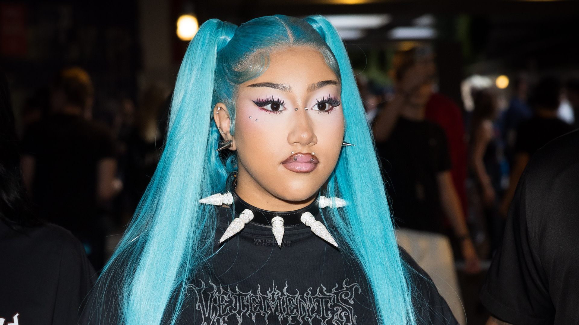 North West with vibrant blue hair in pigtails, wearing a black shirt with white spikes and distinctive makeup, in a crowded setting.