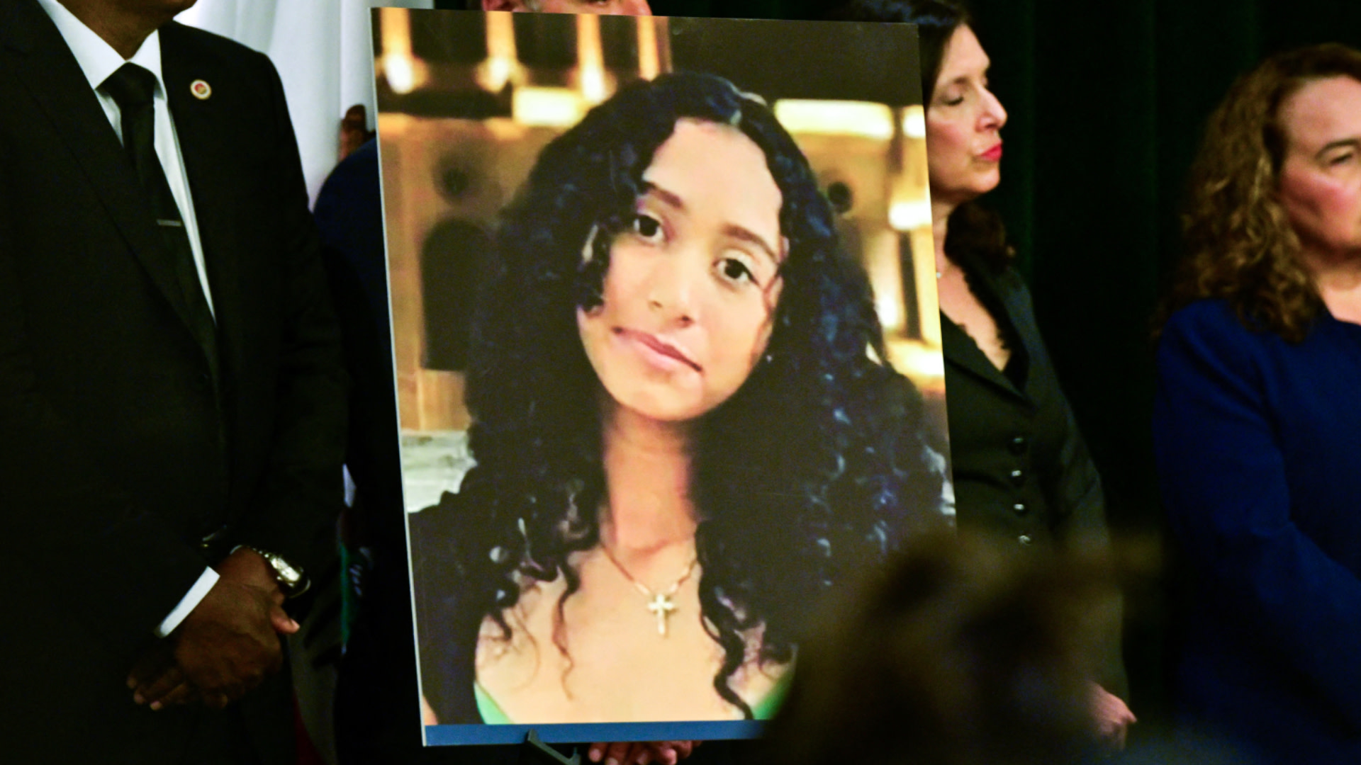 A large photograph of a Celeste Rivas Hernandez with curly hair and a cross necklace is held by people in formal attire.