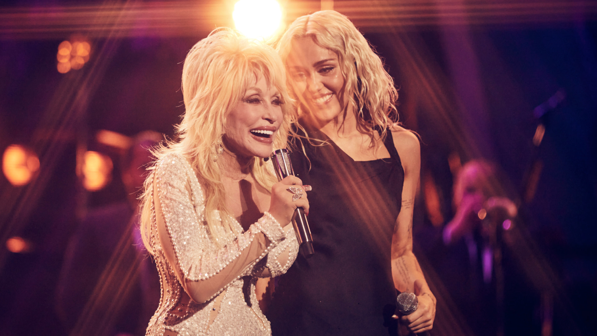 Dolly Parton and Miley Cyrus performing on stage, smiling while holding microphones, with bright stage lights in the background.