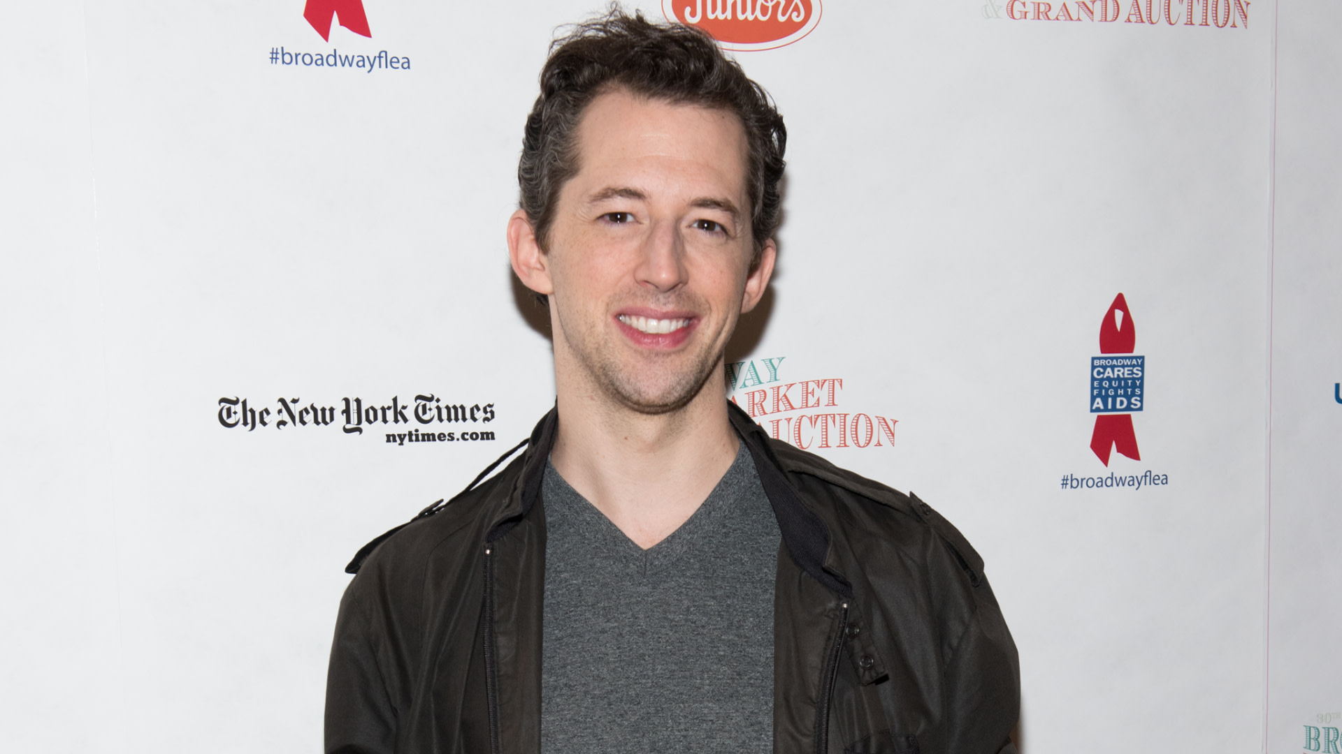 Broadway actor Josh Grisetti in a gray shirt and dark jacket smiles at an event with sponsor logos in the background.