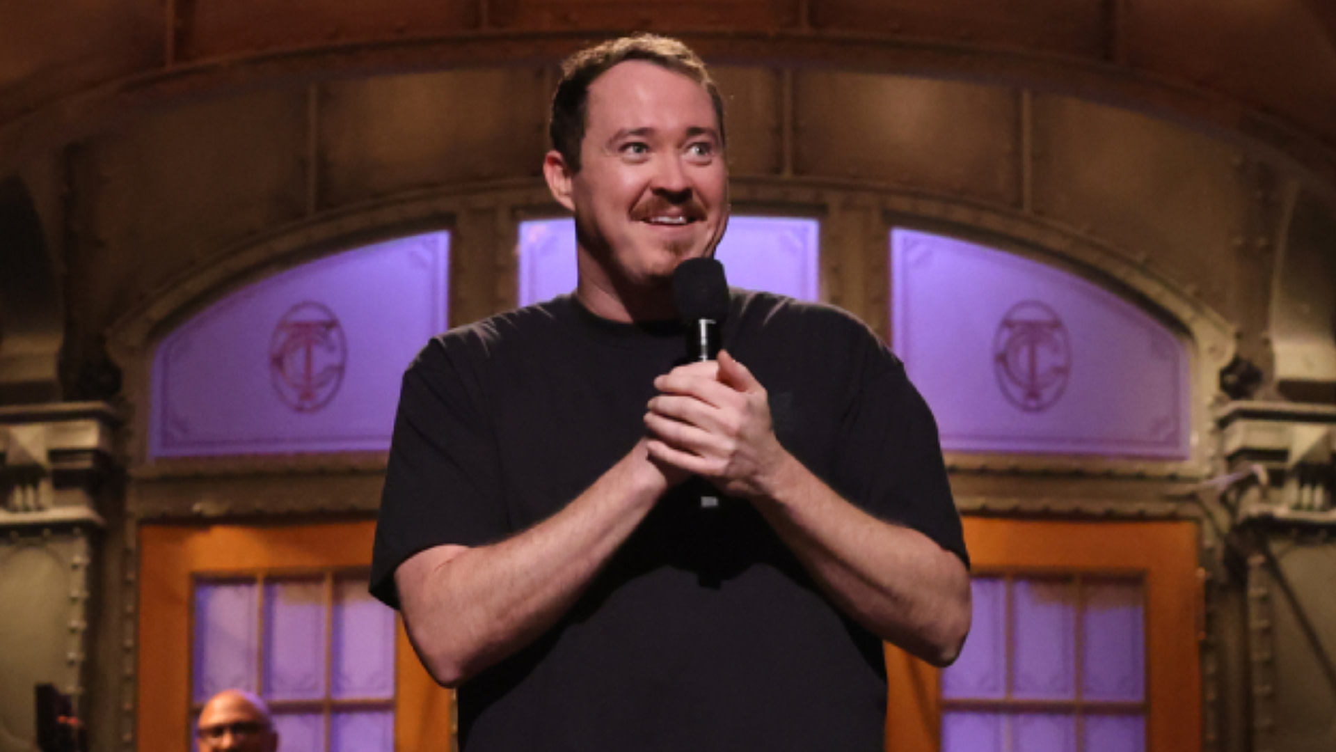 Shane Gillis with short hair and a beard is holding a microphone on stage, smiling. The background features a set with arched windows.