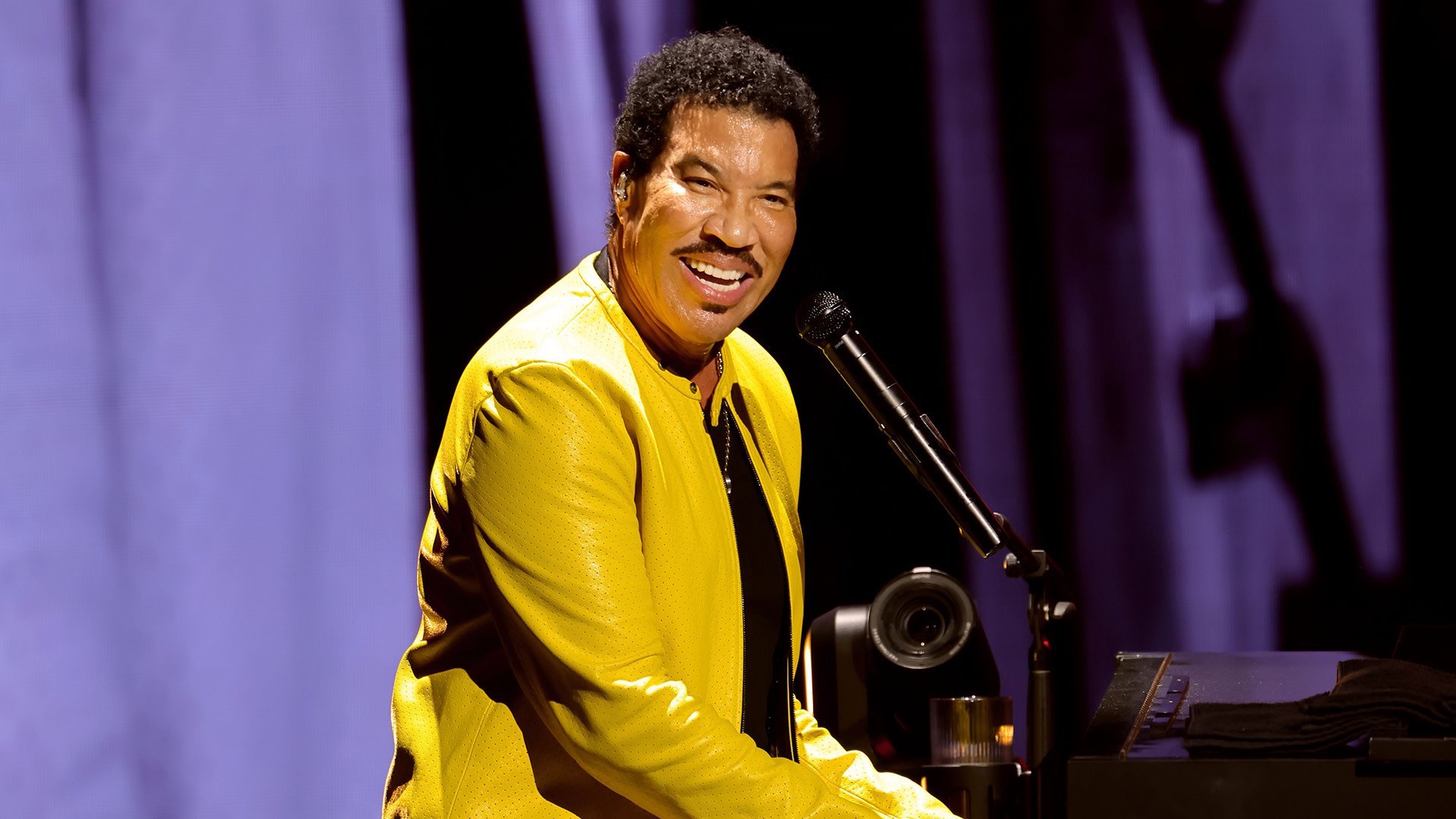 Lionel Richie, wearing a bright yellow jacket, smiles while playing the piano on stage.