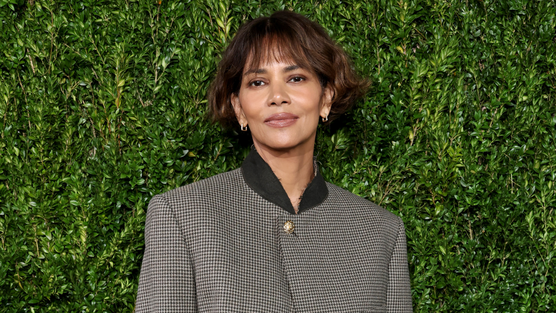 Halle Berry in a checkered jacket, standing in front of a green foliage background.