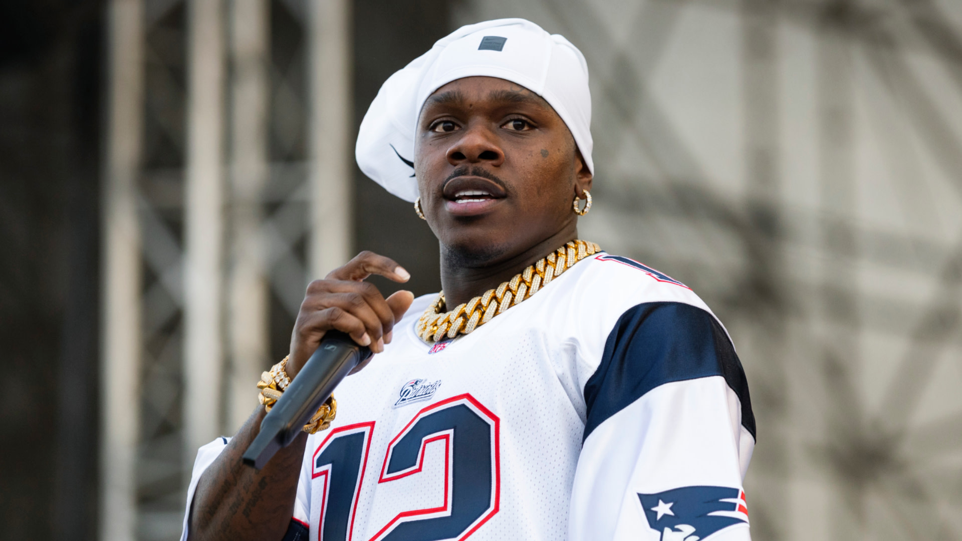 DaBaby holds a microphone on stage. He is wearing a white cap, gold chains, and a Patriots jersey.