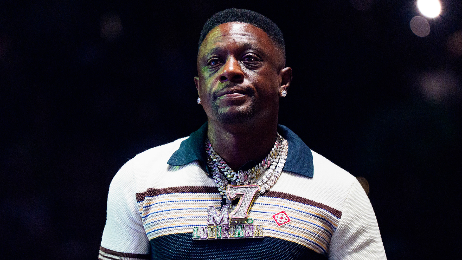 Boosie Badazz wearing a striped shirt and large chain necklace with a pendant, standing against a dark background.