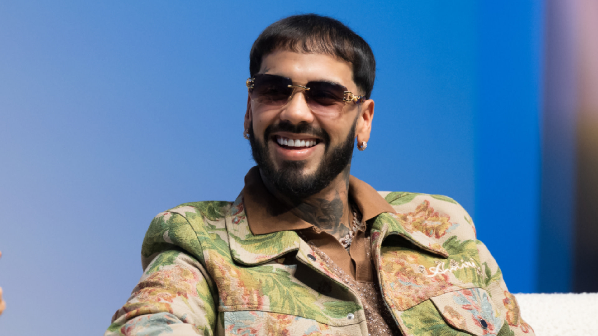 Anuel AA with a beard and sunglasses, wearing a floral patterned jacket, smiles against a blue background.